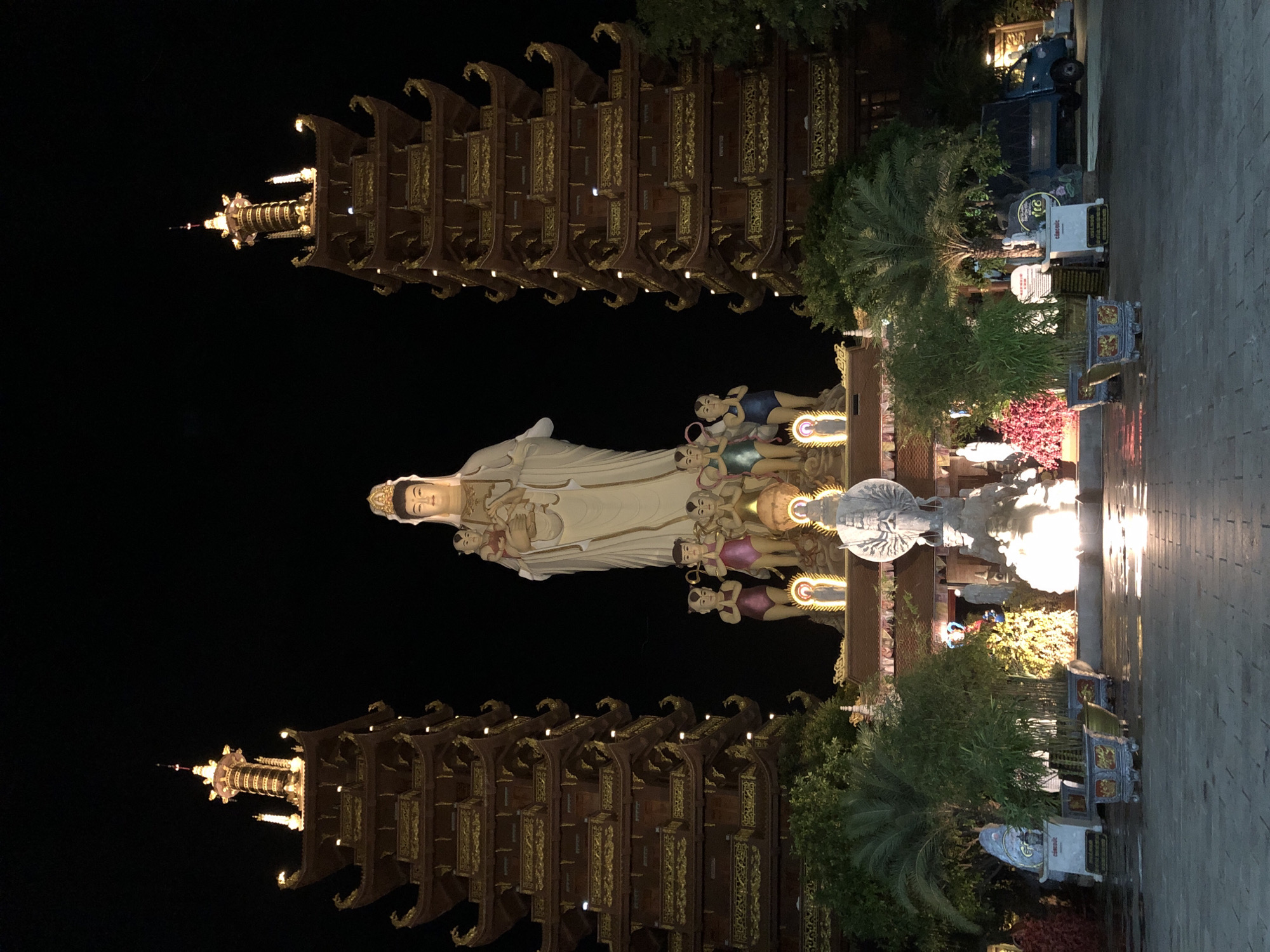 This is a night-time, eye-level shot of the Linh Phong Buddha statue and pagoda in Mũi Né, Vietnam. The central subject is a large, white statue of a woman holding a child, likely a Buddhist deity, with smaller statues of children in front of her. Flanking this central figure are two tall, multi-tiered pagodas, illuminated and intricately decorated with gold accents. The pagodas have a distinctly East Asian architectural style, with curved roofs and ornate carvings. In the foreground, there is a paved courtyard with several potted plants and some decorative lighting. A truck is visible parked to the right, suggesting a public or accessible area. The overall scene is grand and illuminated, emphasizing the religious and architectural significance of the site.