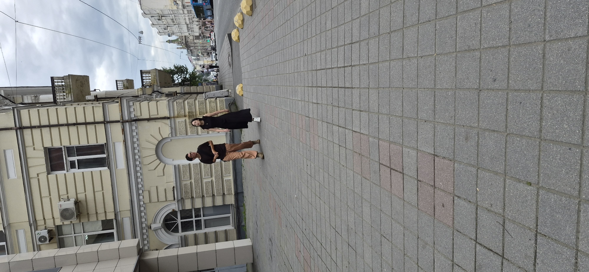 Two people, a man and a woman, walk on a sidewalk. The man, on the left, wears glasses, a dark t-shirt, and light brown pants. The woman, on the right, wears a long black dress and white sneakers. They are walking away from the camera. Behind them, a light yellow building with ornate architectural details lines the street. The sidewalk is paved with gray square tiles. In the background, a street with traffic is visible, including cars, a bus, and overhead wires. The sky is cloudy. The image appears to be taken in a city environment, potentially Vladivostok, Russia, given the architectural style and presence of urban infrastructure.