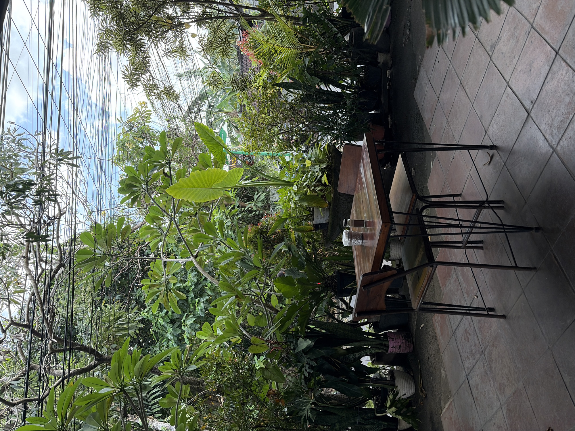 An outdoor seating area is set up with a wooden table and two chairs on a tiled patio. The table is rectangular and appears to be made of dark wood, with two metal chairs with wooden seats placed at opposite sides. A salt and pepper shaker are on the table. The area is surrounded by lush greenery, with various potted plants and trees creating a dense, tropical atmosphere. Overhead, a tangle of electrical wires stretches across the sky, partially obscuring the view of clouds. The scene appears to be in North Kuta, Indonesia, suggesting a cafe or restaurant setting in a tropical climate.