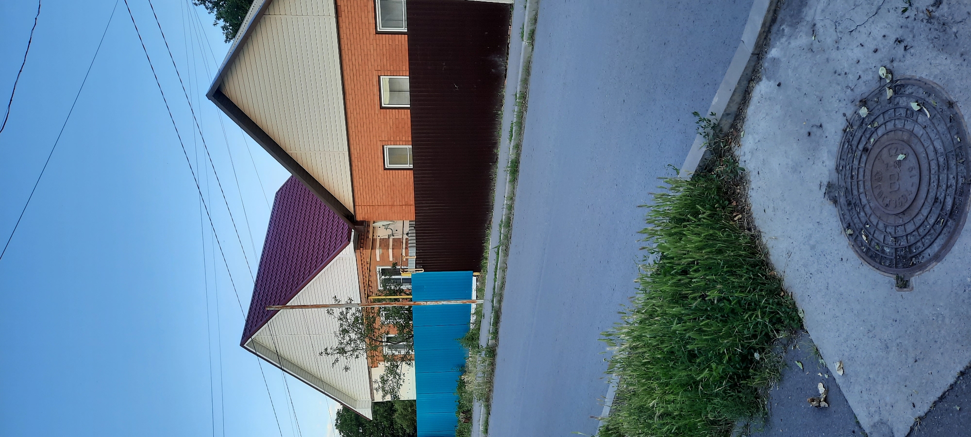 The image depicts a residential street scene in the city of Shakhty, Russia. Two houses with pitched roofs are visible, one with a dark red roof and white siding, and the other with a light beige roof and orange brick facade. Both houses are partially obscured by fences; a bright blue metal fence runs in front of the first house, and a dark brown metal fence separates the second house from the street. The foreground shows a paved road and a sidewalk with green grass and weeds growing along the edge. A circular metal manhole cover is visible on the sidewalk. The sky is clear and blue, suggesting daytime. No people or vehicles are present in the frame.