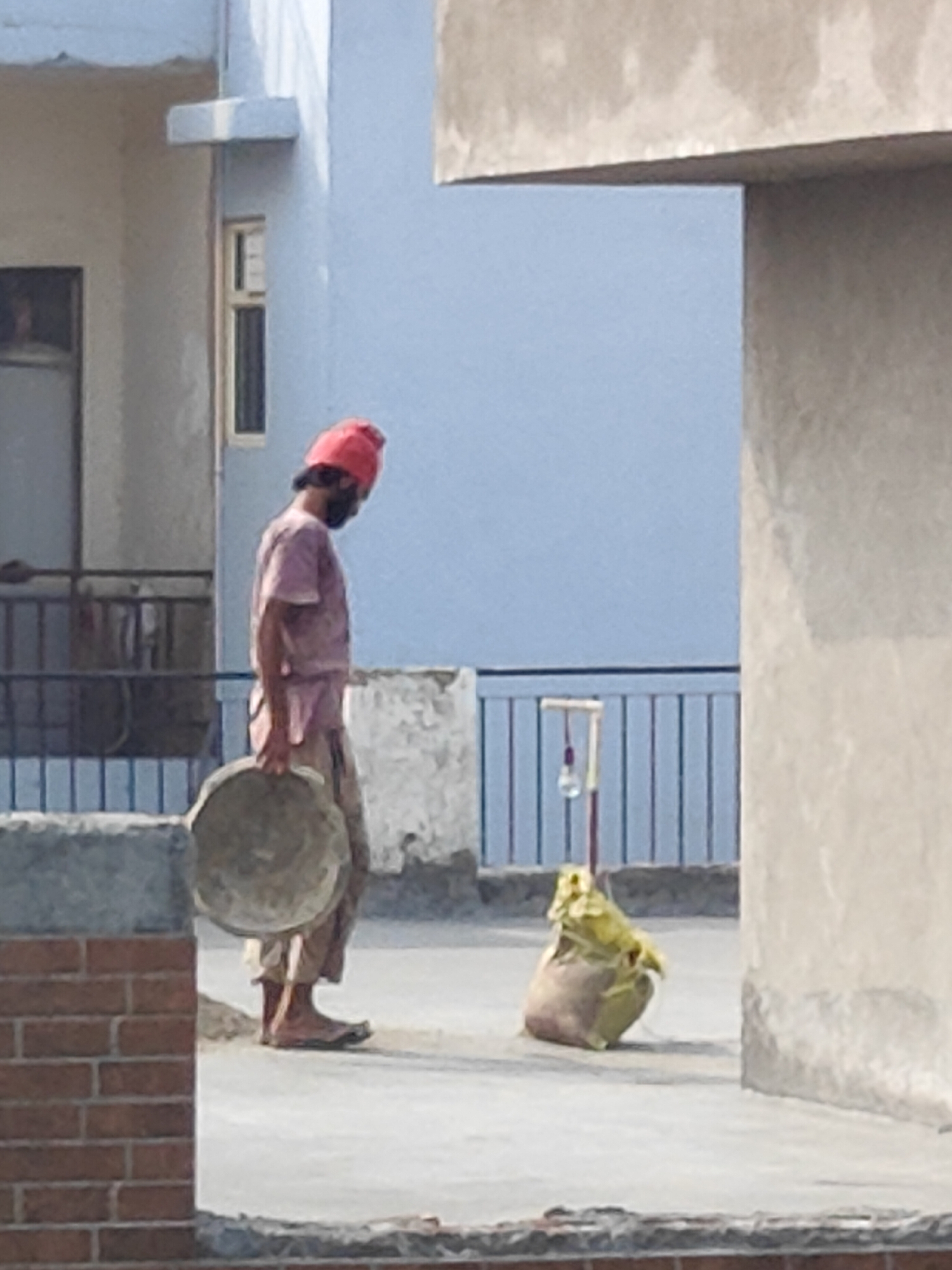 In Gurgaon, India, a lone construction worker, wearing a red turban and a dusty pink shirt and trousers, is captured in a profile view on a rooftop. He is carrying a large, round, grey basin in his right hand, and appears to be walking. Beside him, to his right, is a light brown sack, partially covered with a yellow tarp, resting on the ground. The background features a pale blue building with a window, and further behind, a dark metal railing. The rooftop surface is a light grey concrete. A brick wall is visible in the lower left corner of the frame.