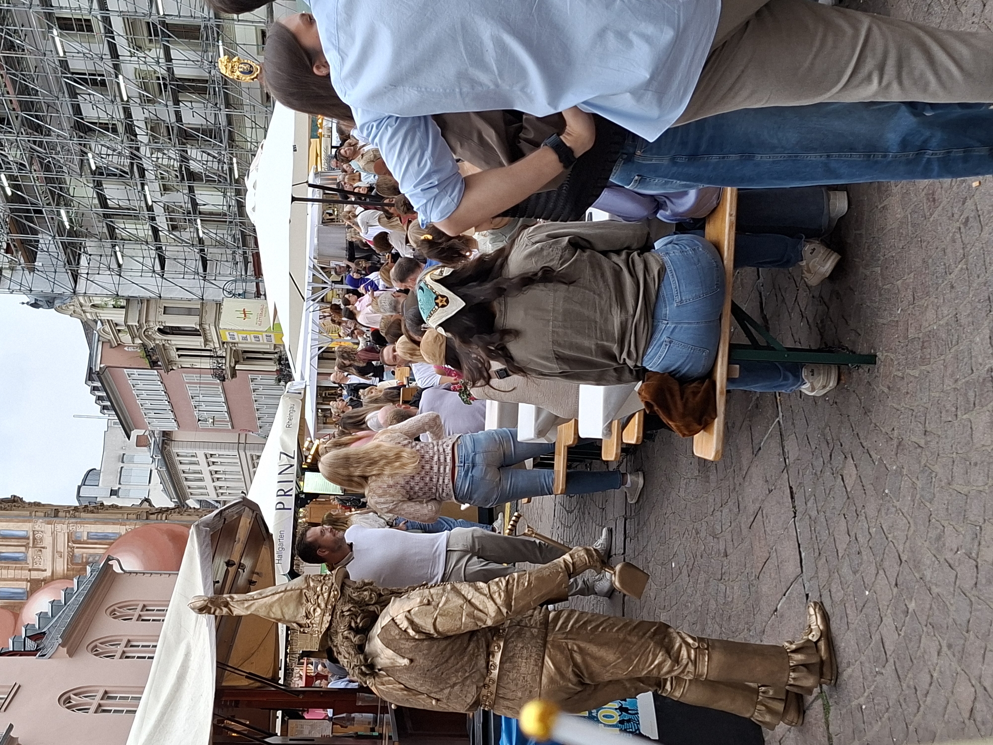 A man dressed in a gold costume, complete with a conical hat and ruffled cuffs, stands on a cobblestone street in Wiesbaden, Germany, holding a small shovel and a golden ball. He appears to be a street performer interacting with a crowd seated at outdoor tables. The crowd is diverse, with men, women, and children enjoying refreshments under white umbrellas and awnings. In the background, buildings adorned with scaffolding suggest ongoing construction or renovation. The overall scene depicts a lively outdoor event or market day in a European city.