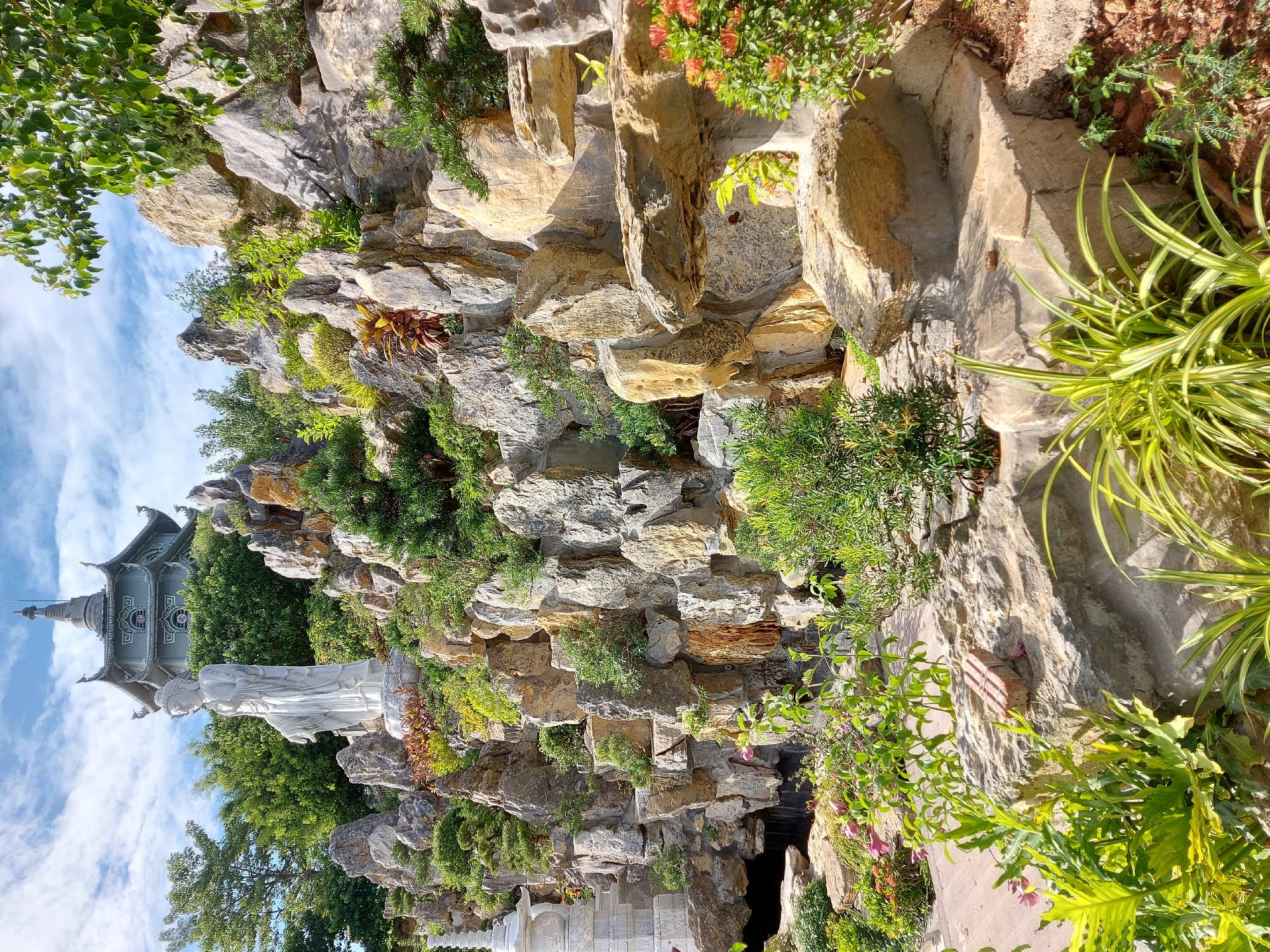 This is a portrait-oriented, eye-level outdoor shot of a lush, rocky garden featuring a large white statue of a figure in flowing robes and an ornate, multi-tiered building with a pagoda-like roof in the background. The statue and building are situated against a backdrop of large, stacked natural rocks, some of which are weathered and have a golden hue. Various green plants, including leafy bushes and spiky foliage, are interspersed among the rocks and at the base of the statue. Some smaller pink flowers are also visible. The sky, visible in the upper left portion of the image, is blue with scattered white clouds. The lighting suggests it is daytime, likely with bright sunlight casting shadows. The overall scene conveys a sense of tranquility and cultural artistry. The location context is Đà Nẵng, Vietnam. No text is visible in the image.