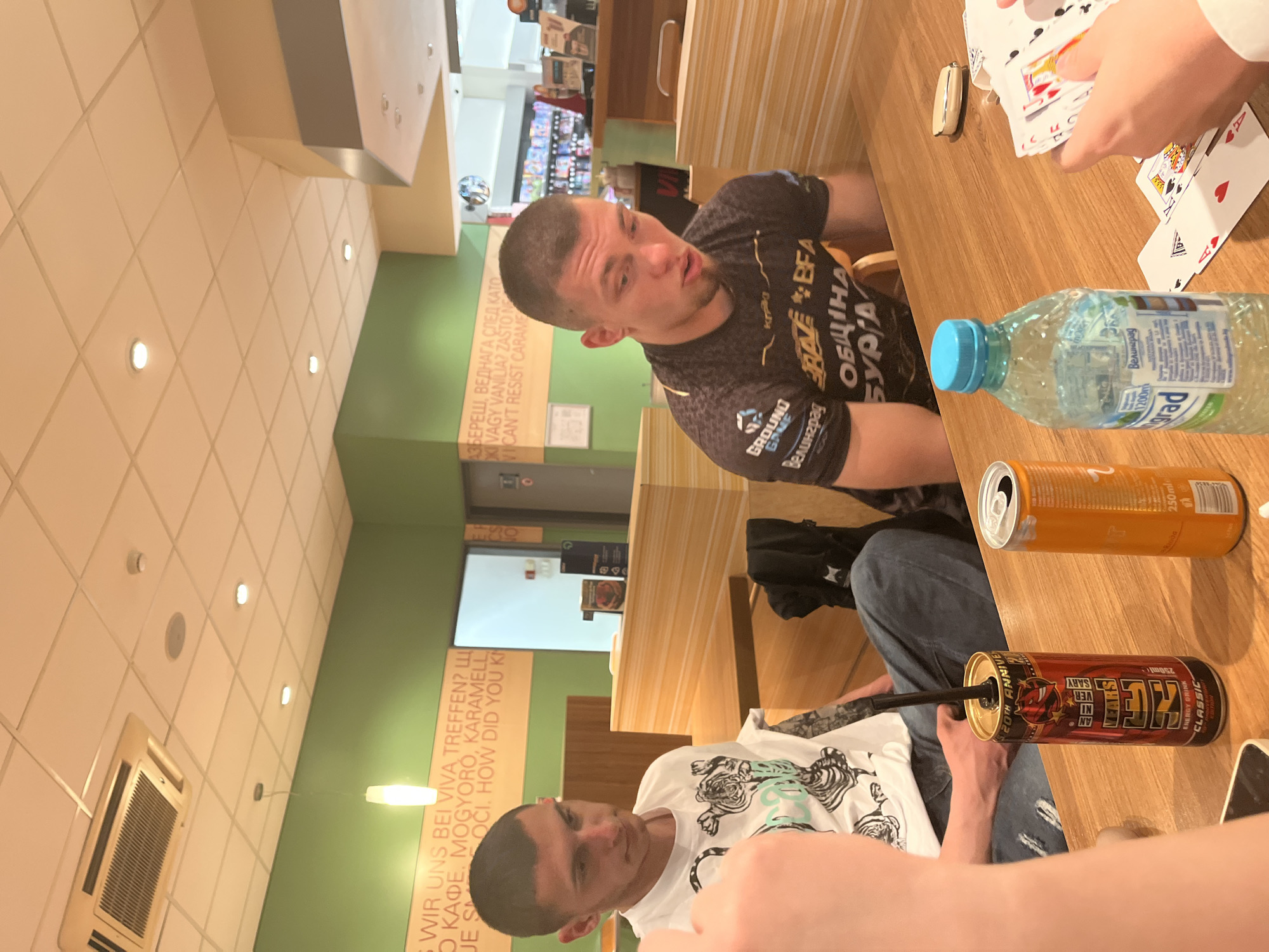 The image captures two men in a casual indoor setting, likely a cafe or fast-food restaurant in Burgas, Bulgaria. Both are seated at a light wooden table. The man on the left, wearing a black t-shirt with logos such as "GROUND GAME," "RAZE BFA," and text in Bulgarian including "Велинград" (Velingrad) and "ОБЩИНА БУРГА" (Municipality Burgas), is looking towards the right with a slightly open mouth, possibly in conversation. He has a short haircut and a light beard.
On the right, another person, largely out of frame, is holding a hand of playing cards, suggesting a card game is in progress. Only their hands and forearms are visible, and the cards include the King of Hearts and an Ace of Spades. Next to the card player, a man in a white t-shirt with a black and teal tiger graphic is partially visible, observing or participating.
On the table are several drinks: a transparent plastic bottle of water labeled "Велинград" (Velingrad) and "1200m" (referring to altitude or volume), an orange energy drink can, and a red "SEVEN KICK" energy drink can with "20TH ANNIVE" and "CLASSIC" visible, from which a black straw protrudes.
The background features light brown and green walls. The green section of the wall displays multi-lingual text, including German phrases like "WIR UNS BEI VIVA TREFFEN?" and "MOGYORÓ, KARAMELL," Bulgarian phrases such as "Щ КАФЕ," "АЗБЕРЕШ, ВЕДНАГА СЛЕД КАТО," and English snippets like "CAN'T RESIST CARAME." The ceiling is tiled in a light, neutral color with embedded recessed lighting and a visible air conditioning unit. The overall atmosphere suggests a relaxed gathering among friends.
