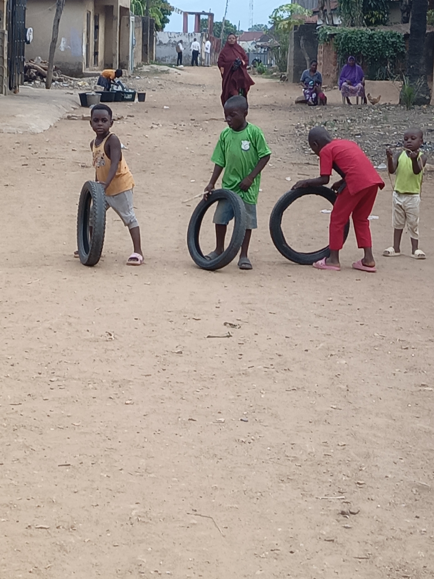 This vertical medium shot captures a vibrant street scene in Akwanga, Nigeria, depicting daily life, likely in the late afternoon.
The primary subjects are four young boys engaged in play on a dusty, light brown dirt road in the foreground. From left to right: a boy in an orange tank top and light pants, wearing pink sandals, looks towards the camera while holding a tire upright. Next to him, another boy in a green t-shirt and grey shorts handles a tire. To their right, a boy in a red t-shirt and red trousers, also in pink sandals, bends over a tire, seemingly preparing to roll it. The smallest boy, wearing a light green t-shirt, light shorts, and white sandals, stands to the far right, holding a stick and observing the tire in front of him. Their activity of rolling tires with sticks is a traditional form of play.
In the midground, the scene expands to include more people and structures. Behind the leftmost boy, a person in an orange top is bending over several dark buckets near a building with a metal gate, possibly performing chores like washing or collecting water. Further down the road on the right, two women are seated outdoors, one in blue and another in purple traditional garments, with a chicken visible nearby. In the background, additional adults can be seen walking or standing, including a man in a white shirt.
The setting is a residential area characterized by simple, somewhat weathered buildings, some with unpainted walls. Patches of green foliage are visible, particularly on the right side of the frame. The sky appears overcast or hazy, suggesting diffuse light rather than direct midday sun. The dry, unpaved road and the general architecture indicate a semi-urban or rural Nigerian community. There is no visible text in the image.