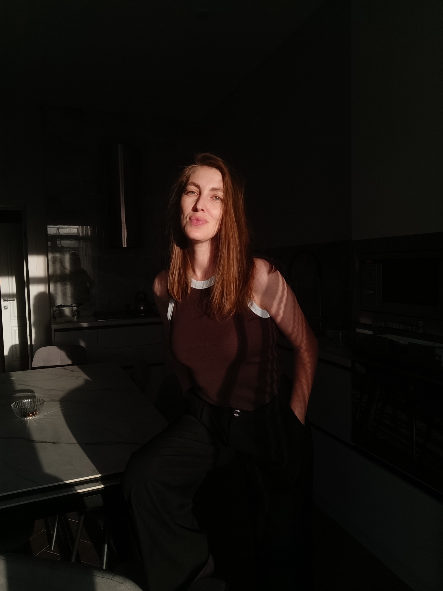 A woman with reddish-brown hair is sitting at a table in what appears to be a kitchen. She is wearing a brown tank top with a white trim and dark pants. Sunlight is streaming in from the left, casting strong shadows across the scene. The woman is looking towards the camera with a slight smile. In the background, kitchen cabinets and appliances are visible, though many areas are in shadow. The table in front of her has a marble top and a small glass bowl containing some small objects. The overall lighting suggests it is either early morning or late afternoon. This scene is located in Chaikovsky, Russia.