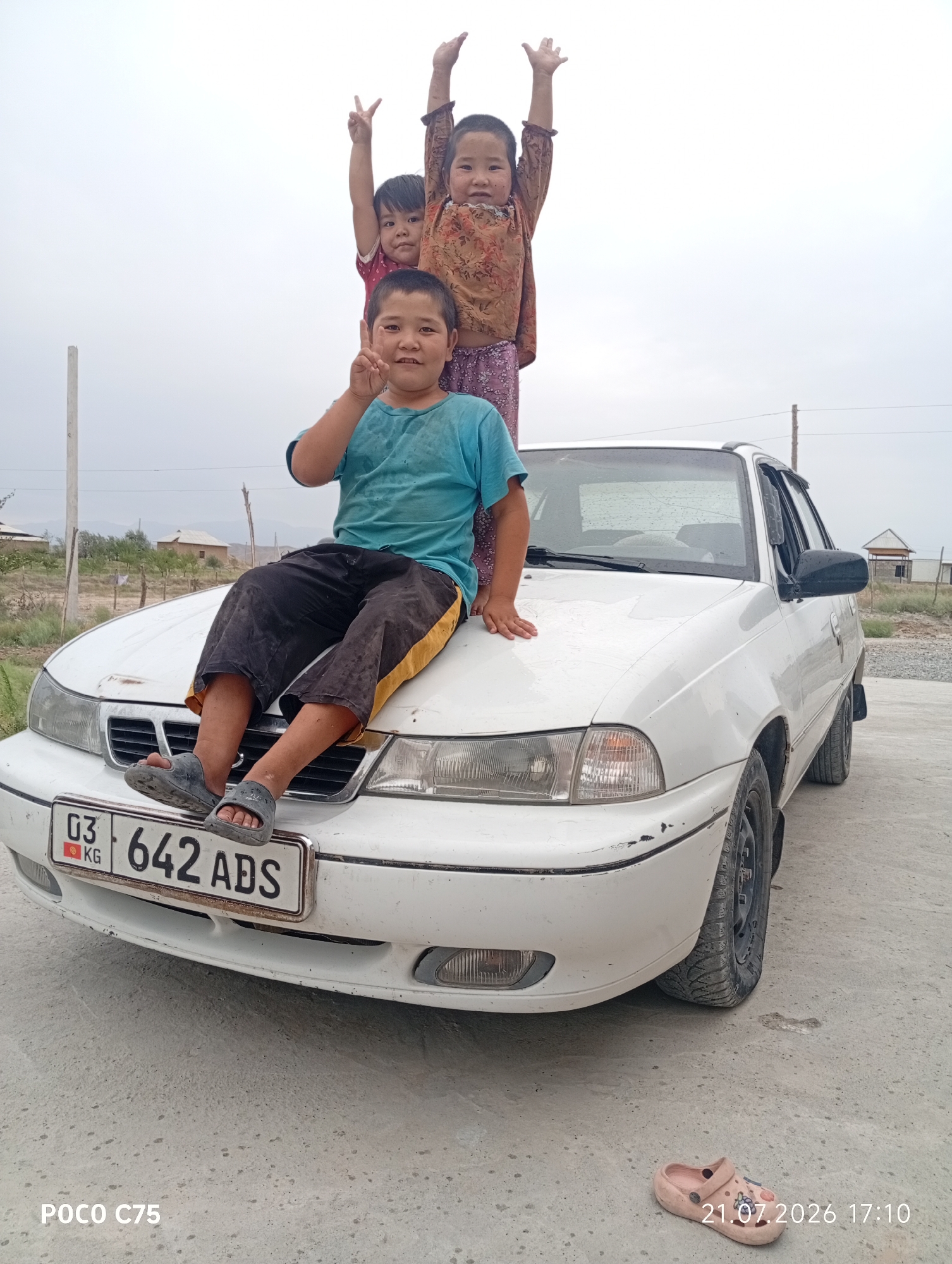 In the village of Chong-Gara, Kyrgyzstan, three children are posing on top of a white sedan. The youngest child, a girl, stands in the middle with her arms raised. To her right, a slightly older girl also has her arms raised. A boy sits on the hood of the car, his legs dangling over the front. He is wearing a blue t-shirt and black pants with a yellow stripe. The car has a license plate that reads "03 642 ADS". The background shows a cloudy sky, some sparse vegetation, and a few buildings. The ground is a dirt and gravel surface.