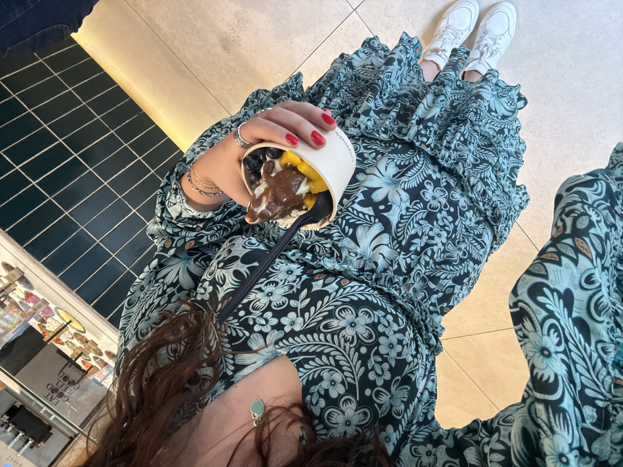 The image is a first-person perspective photograph of a person seated inside a food establishment in Mexico City, Mexico. The person is wearing a floral print dress and white sneakers. They are holding a cup filled with frozen yogurt or ice cream, topped with chocolate sauce, mango chunks, and blueberries. A black spoon is inserted into the cup. The person has red nail polish and is wearing rings and bracelets on their left hand.
In the background, a display case showcasing a variety of food items is visible, alongside a dark blue tiled wall and a beige colored floor. The overall impression is of a casual, indoor setting with a focus on food consumption.