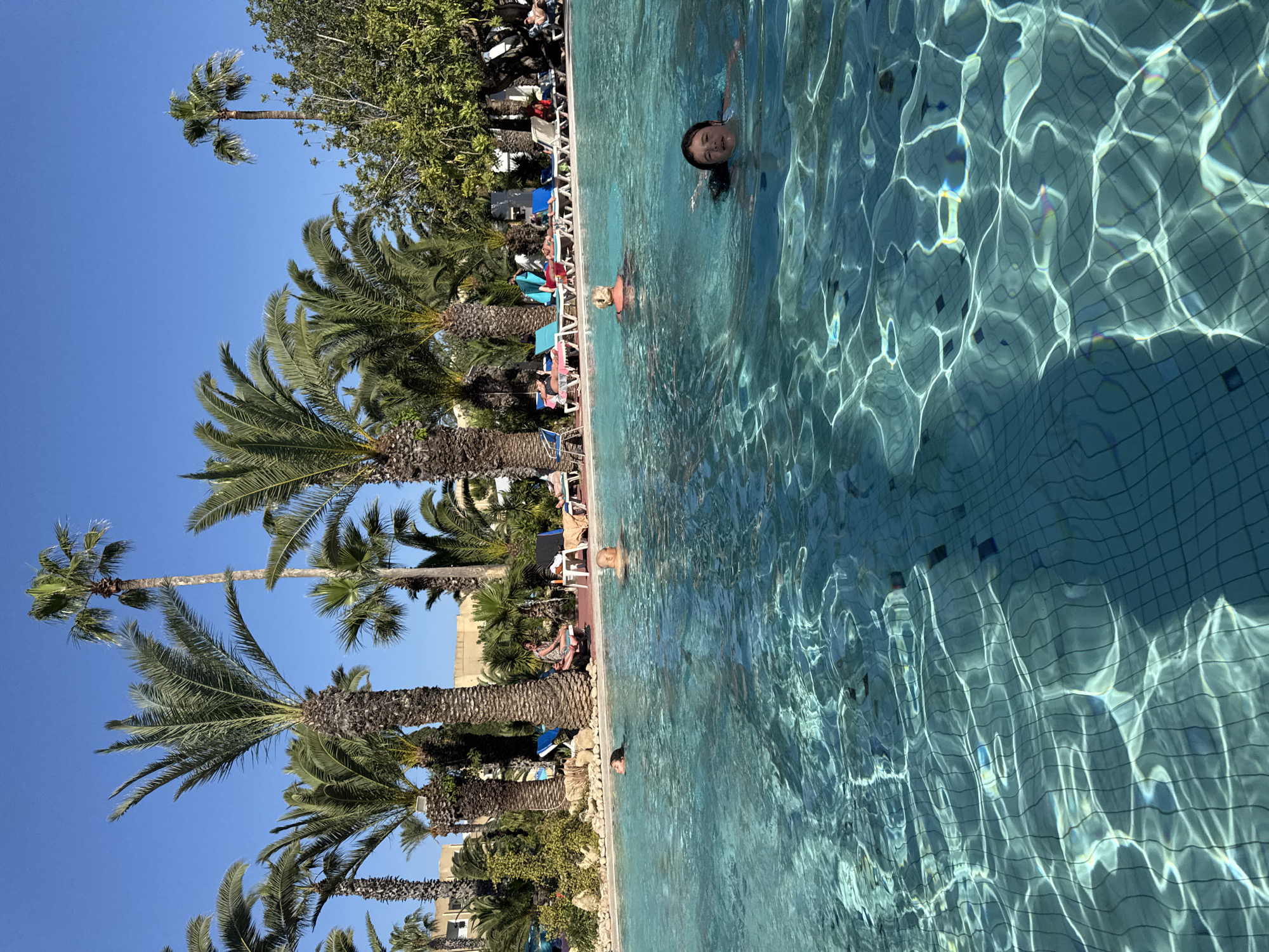 The image shows a swimming pool in Mrezga, Tunisia, surrounded by palm trees and sunbathers. A girl is swimming in the pool, looking directly at the camera. The pool water is light blue, with reflections of sunlight. Lounge chairs and people are visible along the pool's edge. The sky is a clear blue.