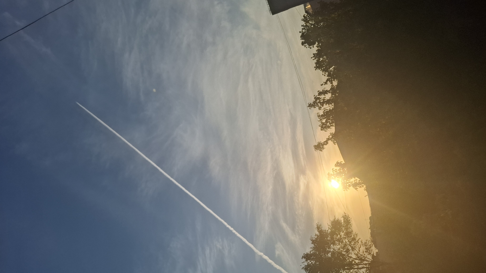 The image captures a sky scene with a contrail from an airplane forming a long diagonal line from the lower left to the upper right. The sky is blue with wispy clouds, and the sun is setting or rising, casting a warm golden glow that illuminates the clouds and trees in the foreground. The lower portion of the image is dominated by silhouetted trees and the rooftops of buildings. A power line is visible stretching horizontally across the upper right portion of the frame. The setting suggests an urban or suburban environment, possibly in Shymkent, Kazakhstan, given the context. There are no people or explicit actions depicted in the image. The overall mood is serene, characterized by the natural light and the vastness of the sky.