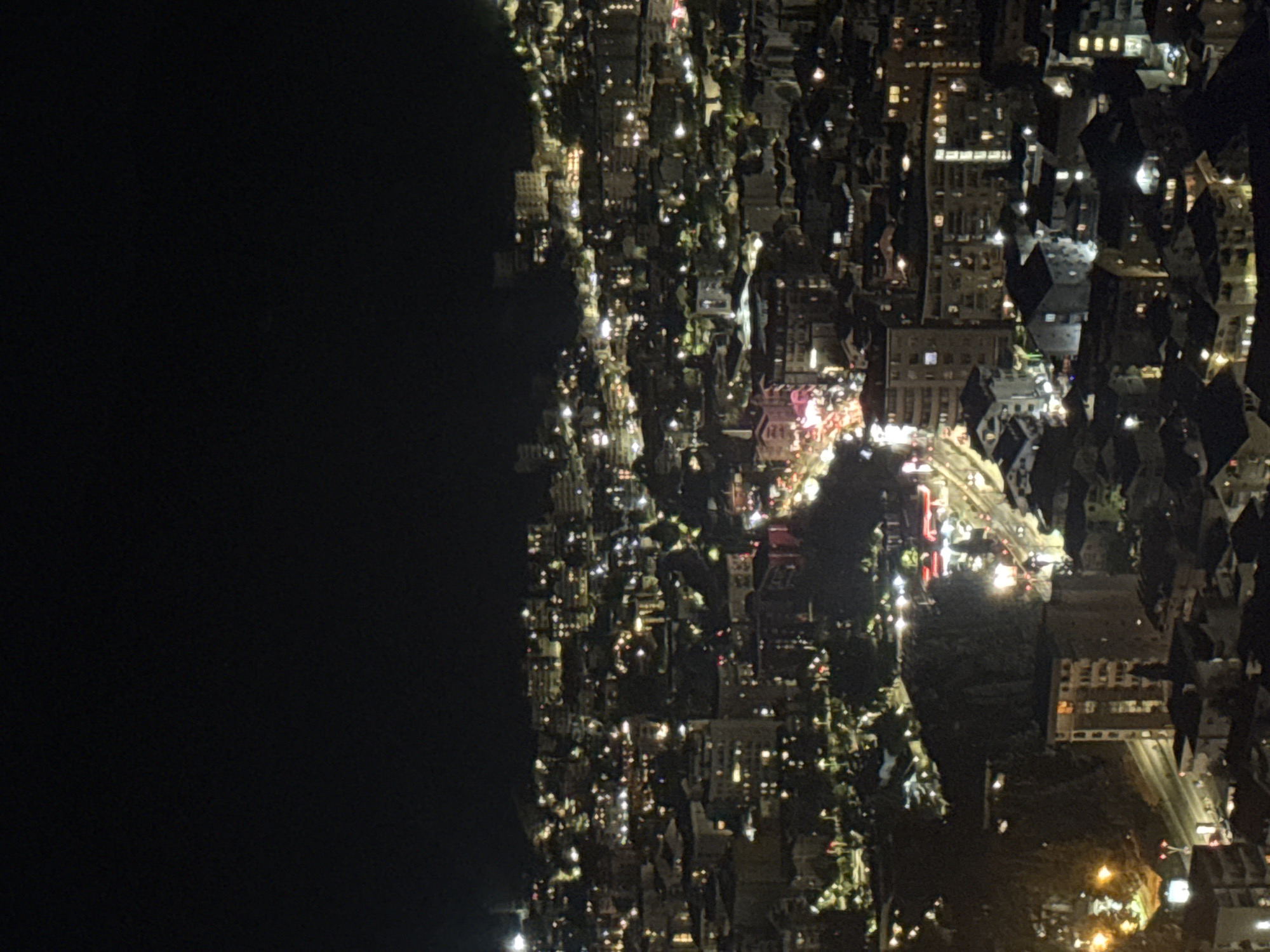 The image is a nighttime, high-angle, wide shot of a densely populated urban area with numerous buildings illuminated by interior lights and streetlights. The cityscape appears to be situated on a hillside, with structures built at varying elevations. The overall scene is dark, with the exception of the artificial lights. In the foreground, a few buildings are clearly visible, including one prominent multi-story structure with illuminated windows. Further into the scene, the lights create a textured mosaic across the landscape, highlighting the density of the urban development. A road with visible lights, presumably from vehicles, snakes through the middle ground of the image. The background fades into darkness, with only distant, faint lights suggesting the continuation of the city or a hazy sky. The image does not contain any people or discernible actions. The environment is exclusively characterized by the built urban infrastructure and the nighttime atmosphere. This image depicts a section of the city of Košljun, Montenegro, at night.
