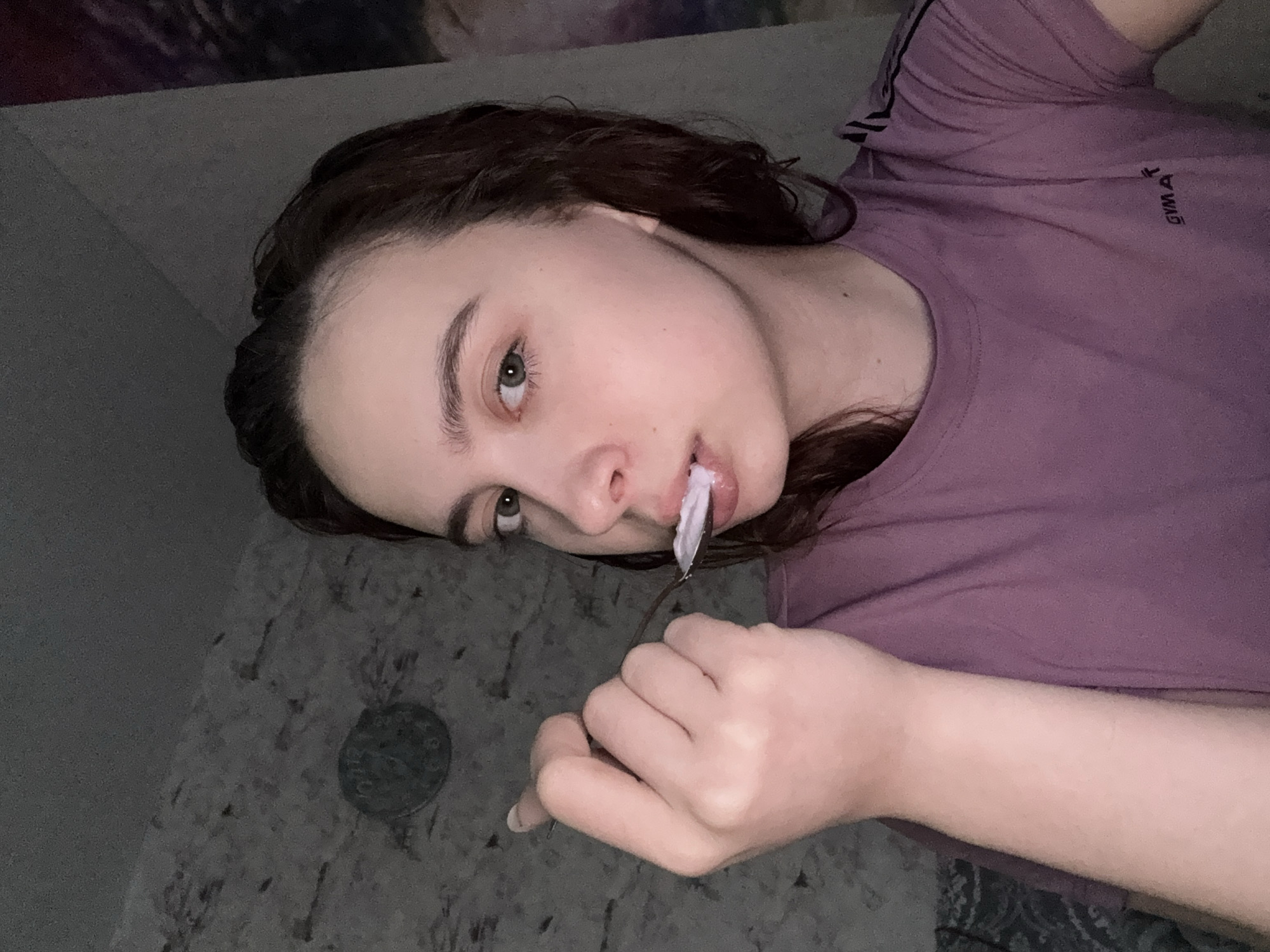 A person with dark, wavy hair and fair skin is lying down, angled towards the camera. They are wearing a purple t-shirt with black stripes on the sleeve and the word "GYMA" printed on the chest. The person holds a fork with a white substance in their mouth. The background appears to be a patterned surface, possibly a counter or wall. A round, dark object is also visible in the background. There are no indications this is in or near Прикамский, Russia.