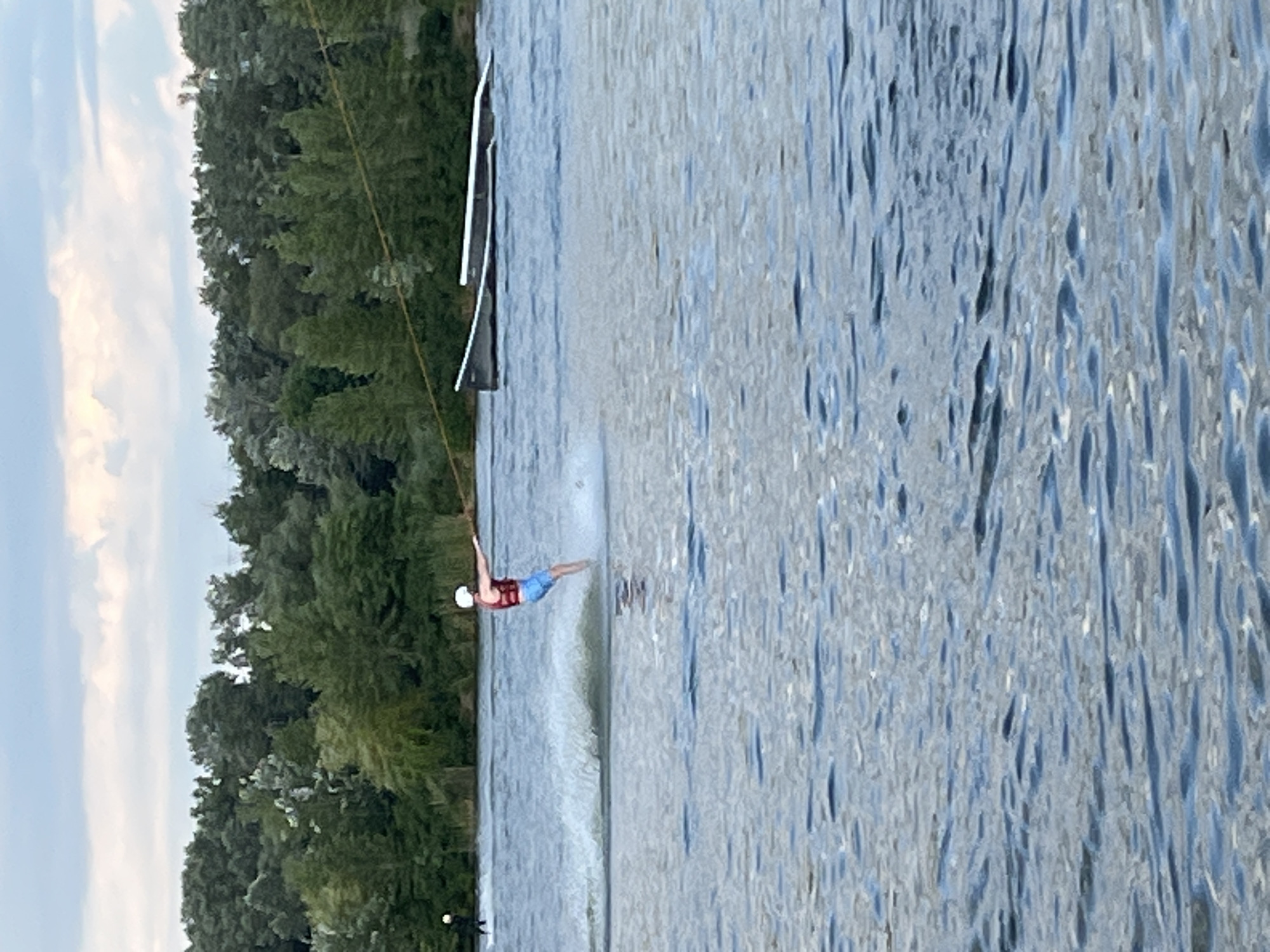 This media file captures a dynamic scene of waterskiing on a lake. The primary subject is a person in mid-action, balanced on skis while being pulled by a tow rope. They are wearing a red life vest and blue shorts, with a white helmet indicating safety precautions. The setting is a body of water bordered by a dense line of lush green trees along the left bank. The water's surface is textured with ripples, suggesting movement and possibly a gentle breeze. In the background, a white structure, likely a ramp or dock, is visible on the water. The sky appears to be overcast with hints of clouds. The overall impression is one of an active outdoor recreational event. The time of day appears to be late afternoon or early evening, given the lighting conditions and the soft, diffused light. While no specific emotional cues are overtly displayed due to the distance and activity, the scene conveys a sense of energetic sport and enjoyment of nature. There is no visible text in the image.