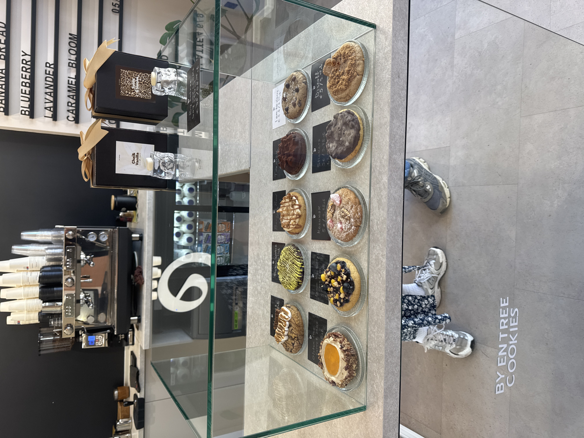 This is an interior shot of a pastry display case inside a bakery, identified as "BY ENTREE COOKIES" and located in Sofia, Bulgaria. The case is filled with an assortment of cookies displayed on individual glass plates, each accompanied by a small sign indicating its name and price. To the left of the display case, a black menu board lists the flavors of other items, including "BANANA BREAD," "BLUEBERRY," "LAVANDER," and "CARAMEL BLOOM." Above this menu, there are two decorative boxes with glass stoppers. A stainless steel espresso machine is also visible on the left. The floor is tiled in a light gray color.
A person is standing in front of the display case, their lower body visible in the reflection of the glass. They are wearing a blue floral dress, white socks, and gray New Balance sneakers. Another person's gray shoe can be seen in the reflection above them.