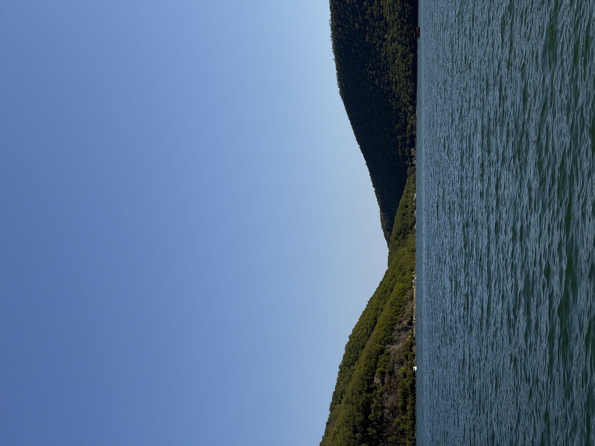 The image captures a portion of Lake Abrau in Abrau-Dyurso, Russia, with a clear blue sky occupying the left side of the frame. The water of the lake, displaying small waves, extends along the right side. On the upper and lower right, forested mountains rise beside the lake.