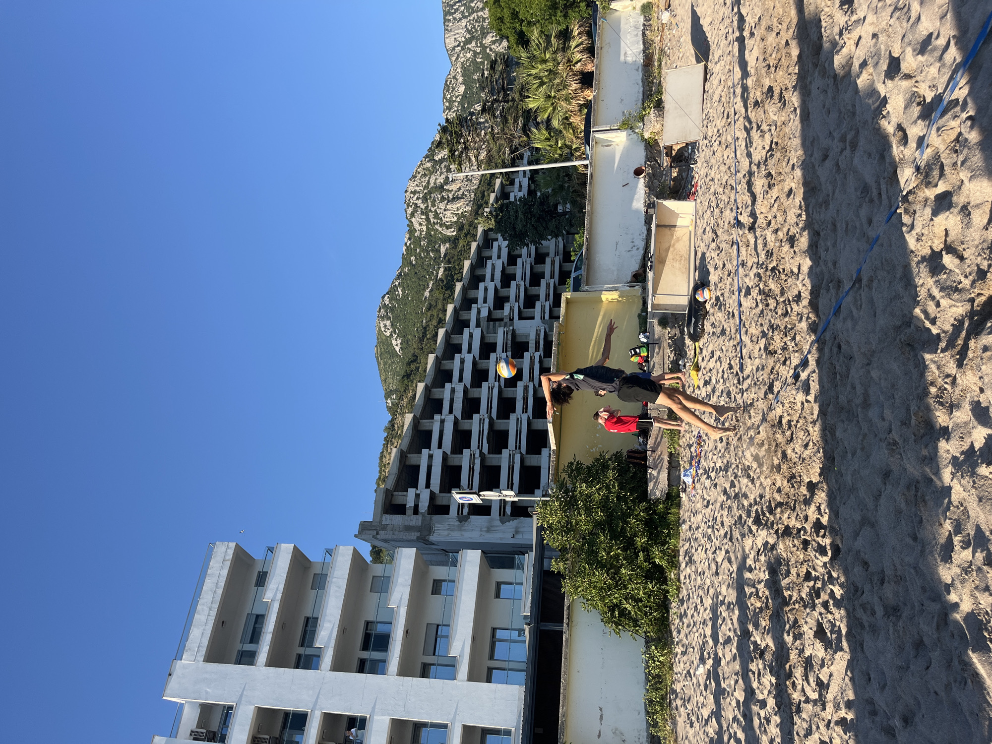 In Zankovići, Montenegro, on a sandy beach under a clear blue sky, a young man in athletic wear is captured mid-air, jumping to hit a volleyball. The ball is visible above and slightly in front of him. Behind the volleyball court, a low, cream-colored wall runs horizontally. Beyond the wall, dense green foliage is visible, including a large bush and several trees, some of which appear to be palm trees. Further in the background, there are two large, multi-story buildings. The building on the left is modern and white with balconies. The building on the right is an unfinished concrete structure with exposed floors and supports, suggesting it is under construction or renovation. Beyond the buildings, a rugged, mountainous landscape with rocky outcrops and sparse vegetation rises steeply, also under the clear blue sky. The sand shows footprints and divots, indicative of activity.