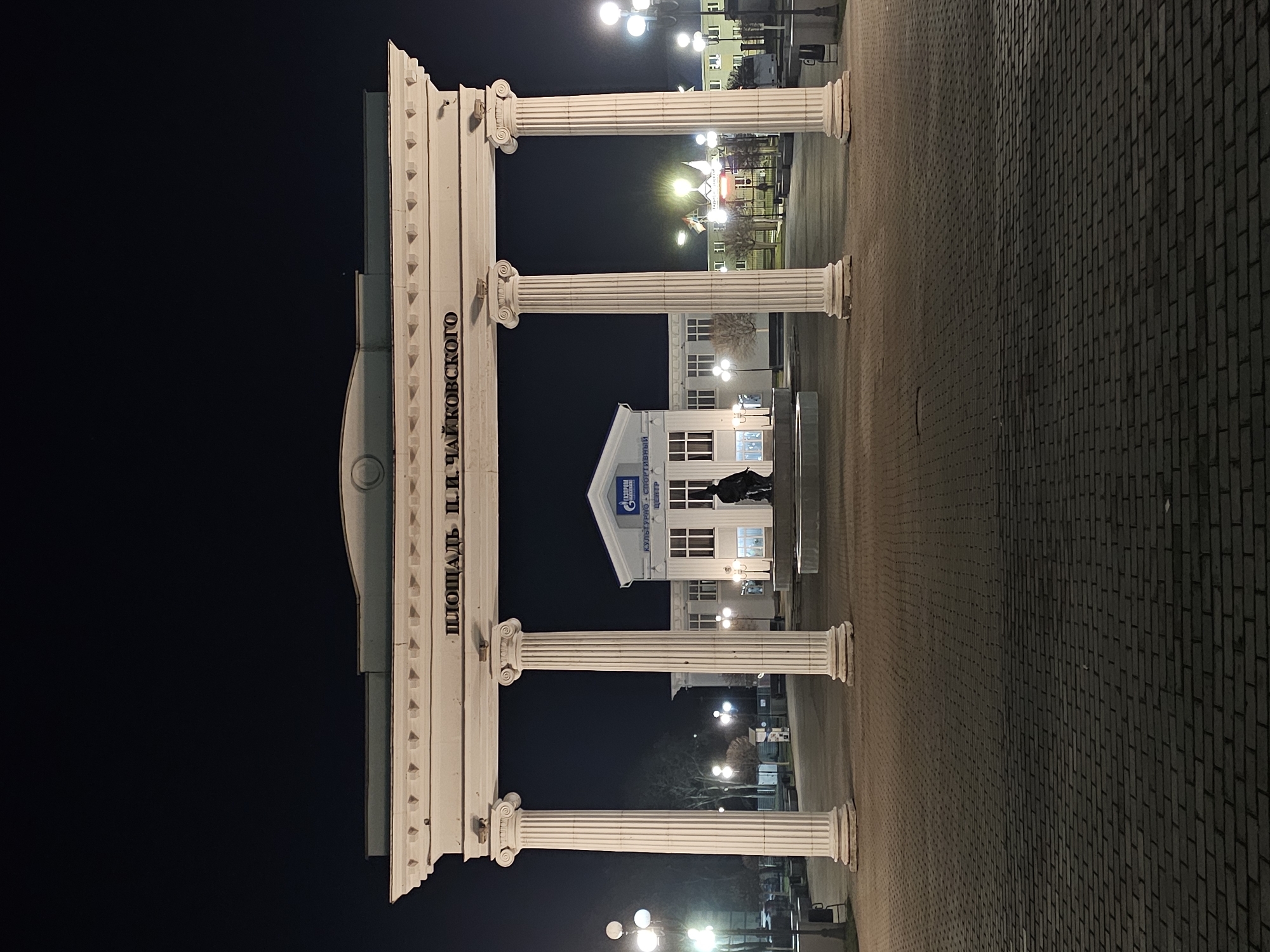 An evening view captures an urban square, likely a plaza, under a dark, clear night sky. In the foreground, a prominent white archway with four classical columns, featuring Ionic capitals, frames the scene. The architrave above the columns bears the Russian text "ПЛОЩАДЬ П.И. ЧАЙКОВСКОГО," which translates to "P.I. Tchaikovsky Square."
Beyond the archway, a brightly lit building with a white facade and numerous windows stands. The roof of this building features a blue logo for "GAZPROM" (Газпром) and the text "КУЛЬТУРНО - СПОРТ ЦЕНТР" (Culture - Sports Center) below it. Partially visible on the left side of the building's facade is "ТОРГОВЫЙ ЦЕНТР" (Shopping Center). In front of this building, a dark statue of a person is visible.
The ground of the square is paved, with lighter-colored paving stones closer to the archway and darker, brick-like paving extending further into the distance on the right. Numerous globe-shaped streetlights illuminate the square, casting warm light and creating reflections on the ground. The overall impression is a well-maintained public space that is active at night. There are no people visible in the image.