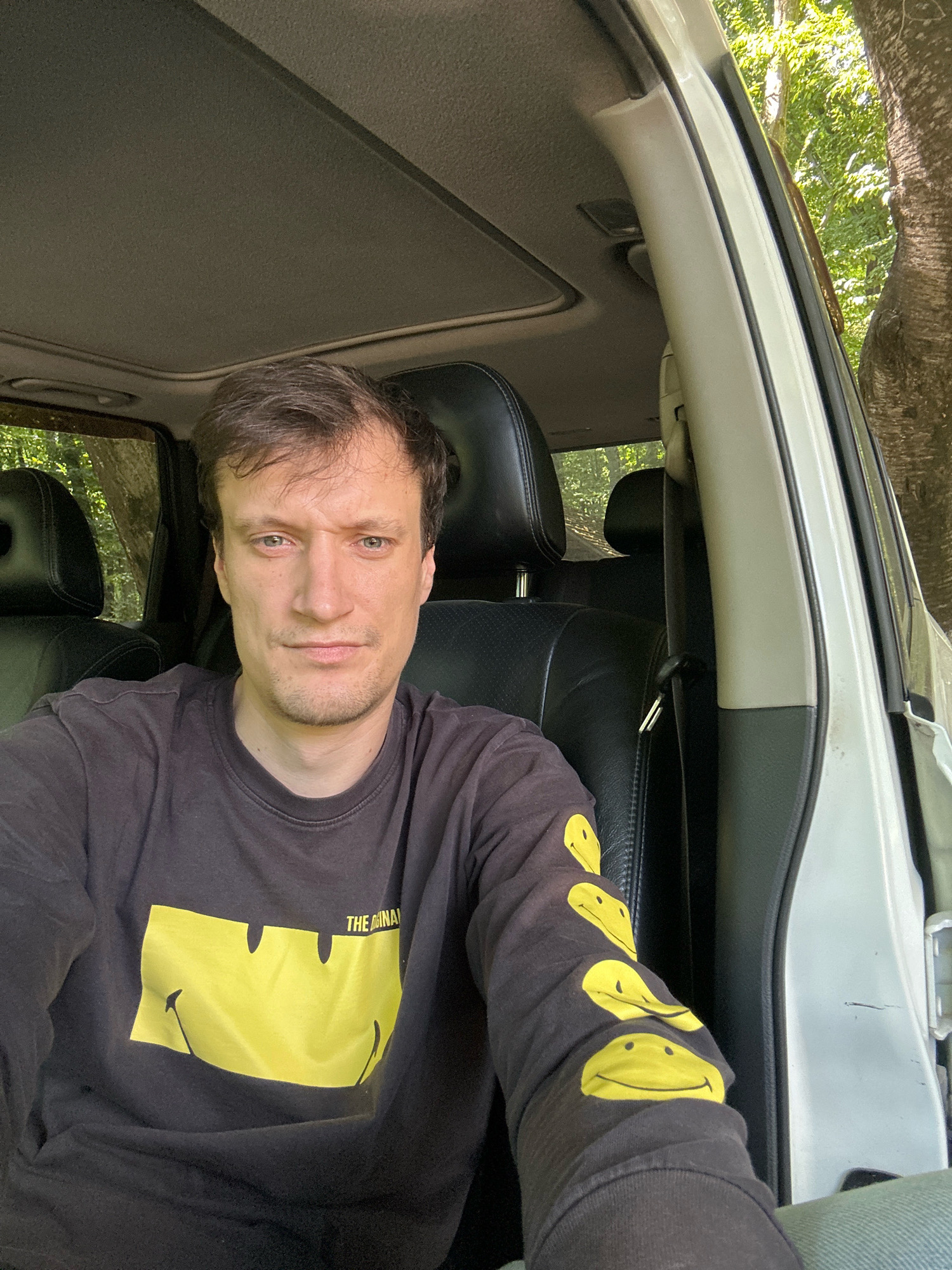 The image is a close-up selfie taken from the driver's seat of a vehicle. The subject, a man with brown hair and fair skin, is looking directly at the camera with a neutral expression. He is wearing a black long-sleeved shirt with a yellow smiley face graphic on the front and several smiley faces on the right sleeve. The interior of the vehicle is visible, including black leather seats, the dashboard, and a sunroof. The exterior of the vehicle shows a white doorframe, and through the open door and windows, a lush green environment with trees is visible. The location is identified as Shatava, Ukraine.
