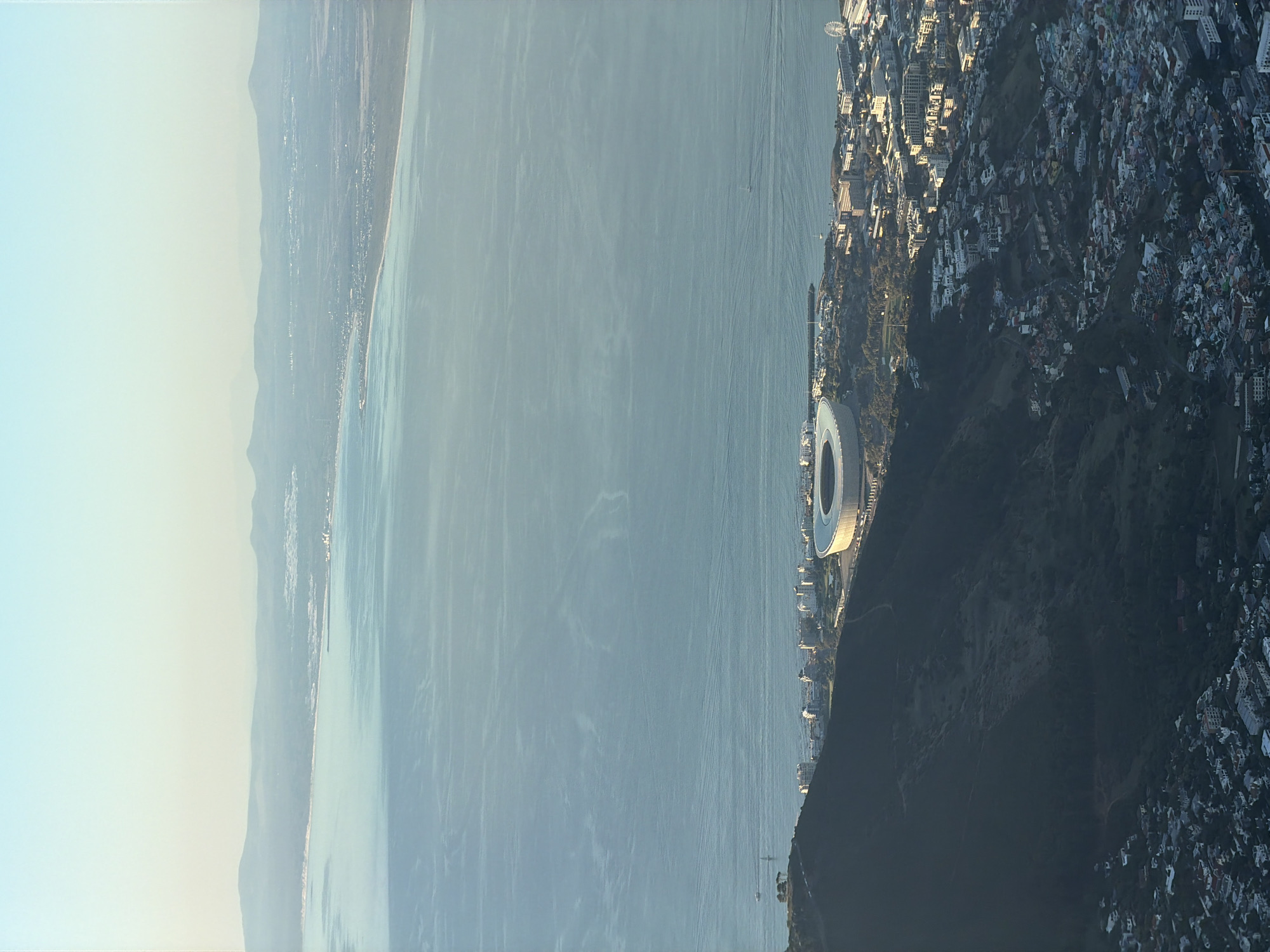 This media file presents an expansive aerial panorama of Cape Town, South Africa, showcasing a coastal city, a vast body of water, and distant mountain ranges. The image is oriented such that the primary urban area and coastline run along the right side, a large expanse of water occupies the middle, and hazy landmasses stretch across the left horizon.
The city on the right is densely packed with buildings, extending from the waterfront up the slopes of hillsides, which are partially covered in dark green vegetation. A prominent landmark is a modern, circular stadium with a distinctive white, somewhat translucent roof and a dark-colored exterior, situated conspicuously near the water's edge. The urban sprawl includes a mix of residential and commercial structures, with varying architectural styles visible.
The central part of the image features a wide, calm body of blue-grey water, likely Table Bay or the Atlantic Ocean. Its surface shows subtle ripple patterns and faint, elongated lines indicating the wakes of distant boats, though the vessels themselves are mostly indistinct due to the distance. A few small, indistinct shapes that could be boats are barely visible on the water.
In the background, on the far left, a series of elongated, distant mountain ranges or hills rise subtly above the water. These landforms appear softened and muted in color, exhibiting atmospheric haze, with some signs of coastal development or smaller settlements along their bases.
The clear sky transitions from a light, pale blue at the top to a warmer, yellowish-white closer to the horizon, suggesting either an early morning or late afternoon light. This soft lighting casts gentle shadows across the urban landscape, highlighting the textures of the stadium and buildings. No specific human activity, such as people, cars, or active events, is clearly discernible, nor is any text visible. The overall scene conveys the grandeur and geographical diversity of the Cape Town area.