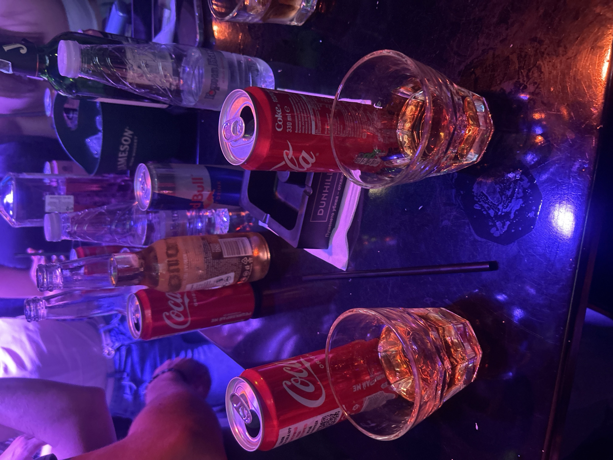 The image presents a cluttered tabletop scene in what appears to be a bar or club. There are multiple bottles and cans scattered around, including brands like Jameson, Red Bull, Coca-Cola, and a bottle of "Gorna Bal". Two partially filled glasses containing amber-colored liquids (likely alcoholic beverages) are positioned near opened Coca-Cola cans. A black drinking straw and a pack of Dunhill cigarettes are also present. The lighting is dim, with both blue and orange hues illuminating the scene, suggesting a nightlife atmosphere. In the background, there are hints of people sitting or standing, with blurred clothing and limbs visible. The image does not give any evidence that the image was taken in Veliko Tarnovo, Bulgaria.