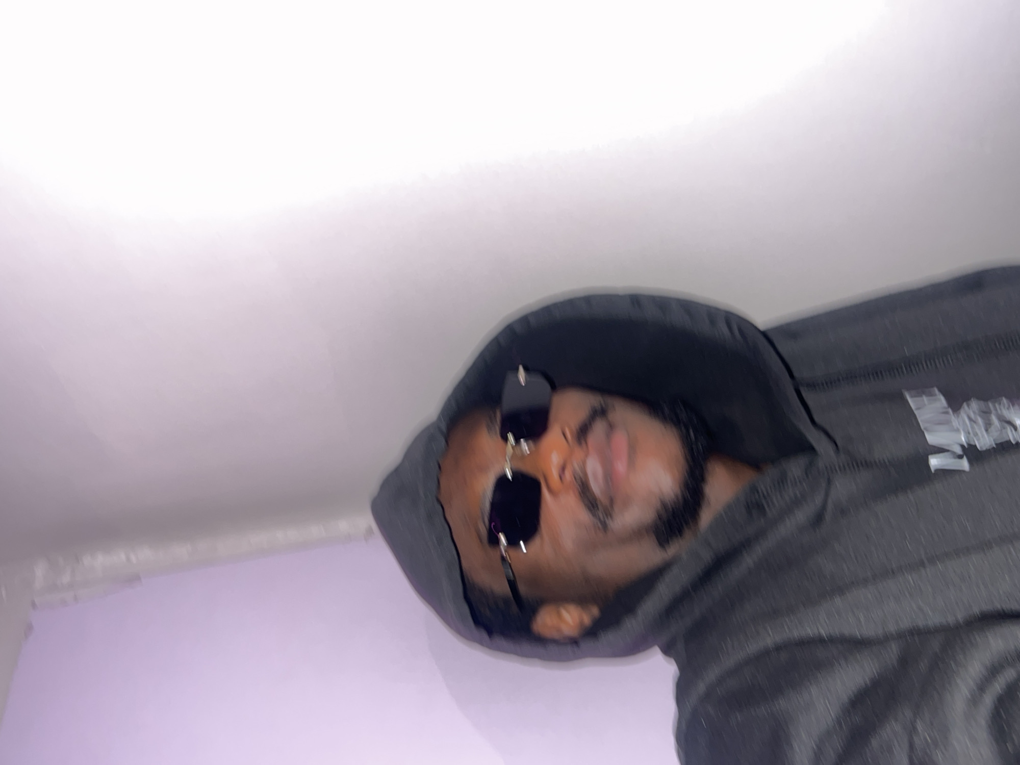 A person wearing sunglasses and a gray hooded sweatshirt is indoors. The background is a white and light purple wall. The location is unknown and cannot be determined from the image.