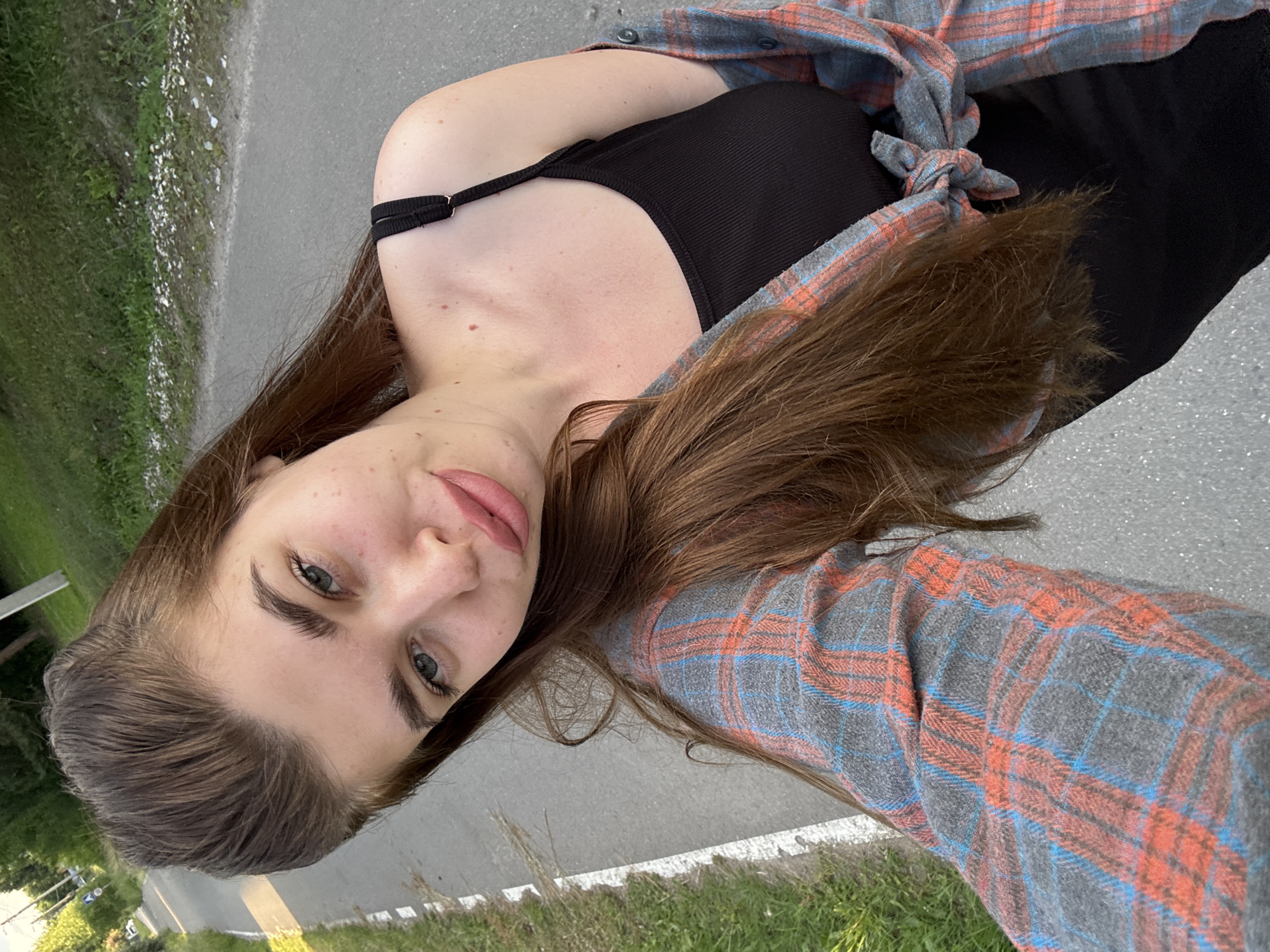 A young woman with long, brown hair and blue eyes is pictured from a high angle, looking directly at the camera. She is wearing a black tank top and an open, grey flannel shirt with orange and blue plaid patterns tied around her waist. Her skin is fair, with some freckles visible. Her lips are slightly parted, and she has a neutral expression.
The setting appears to be outdoors, possibly by the side of a road, with a grassy area and trees visible in the background. The ground is paved, with a white line marking the edge of the road. The lighting suggests it is daytime, with soft natural light. The overall mood is casual and relaxed. No text is visible in the image. The location context of Мещёры, Russia is provided.