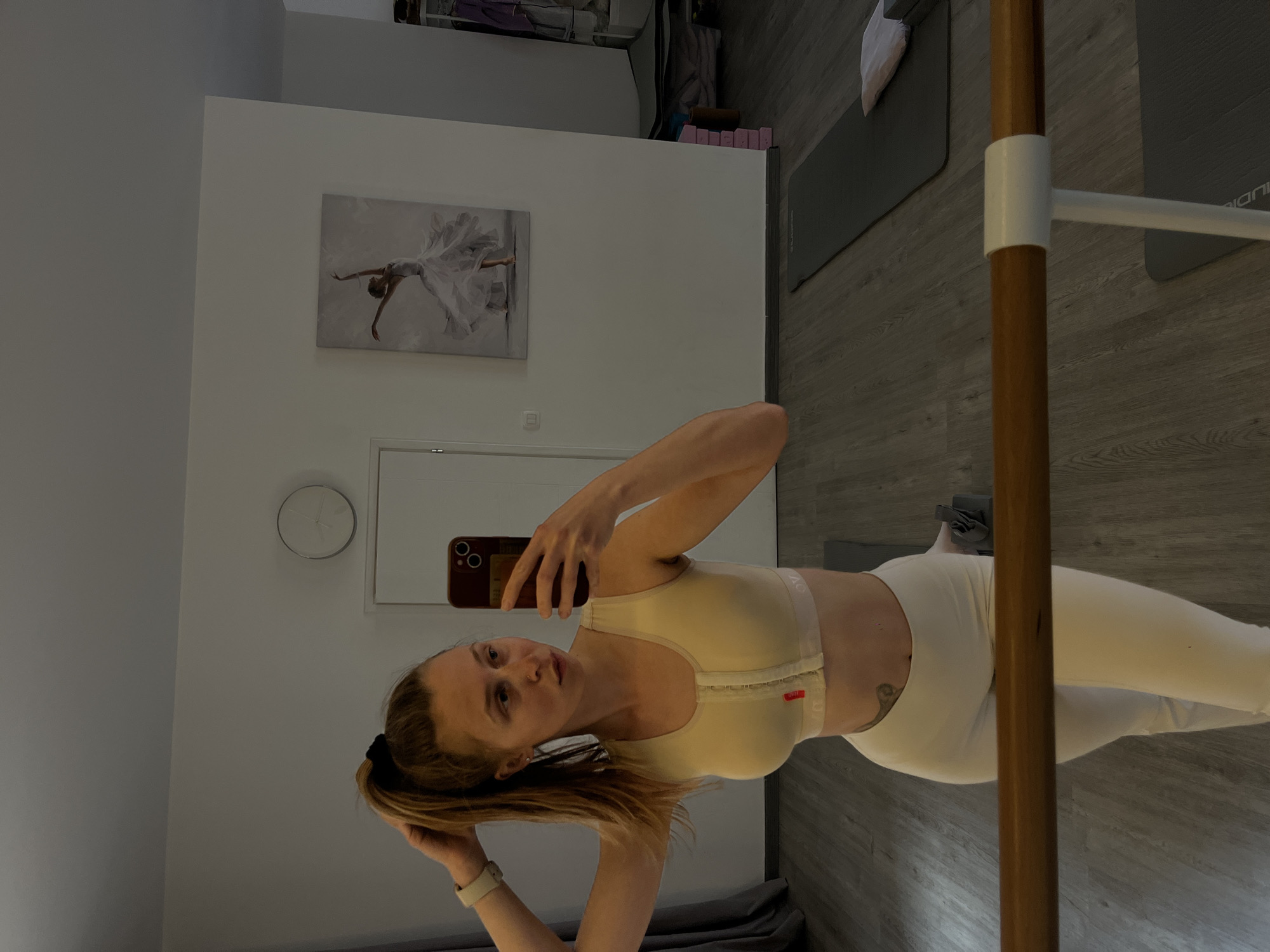 A woman is taking a selfie in front of a mirror, holding a phone in her right hand. She's wearing a beige sports bra with a front zipper and white leggings. Her hair is tied back in a ponytail. Behind her, the scene shows part of a room with a ballet barre. There's a painting of a dancer on the wall, a clock, and yoga mats on the floor. The location is likely indoors, possibly in an apartment or studio in the city of Budva, Montenegro, but there is no evidence to confirm the city.