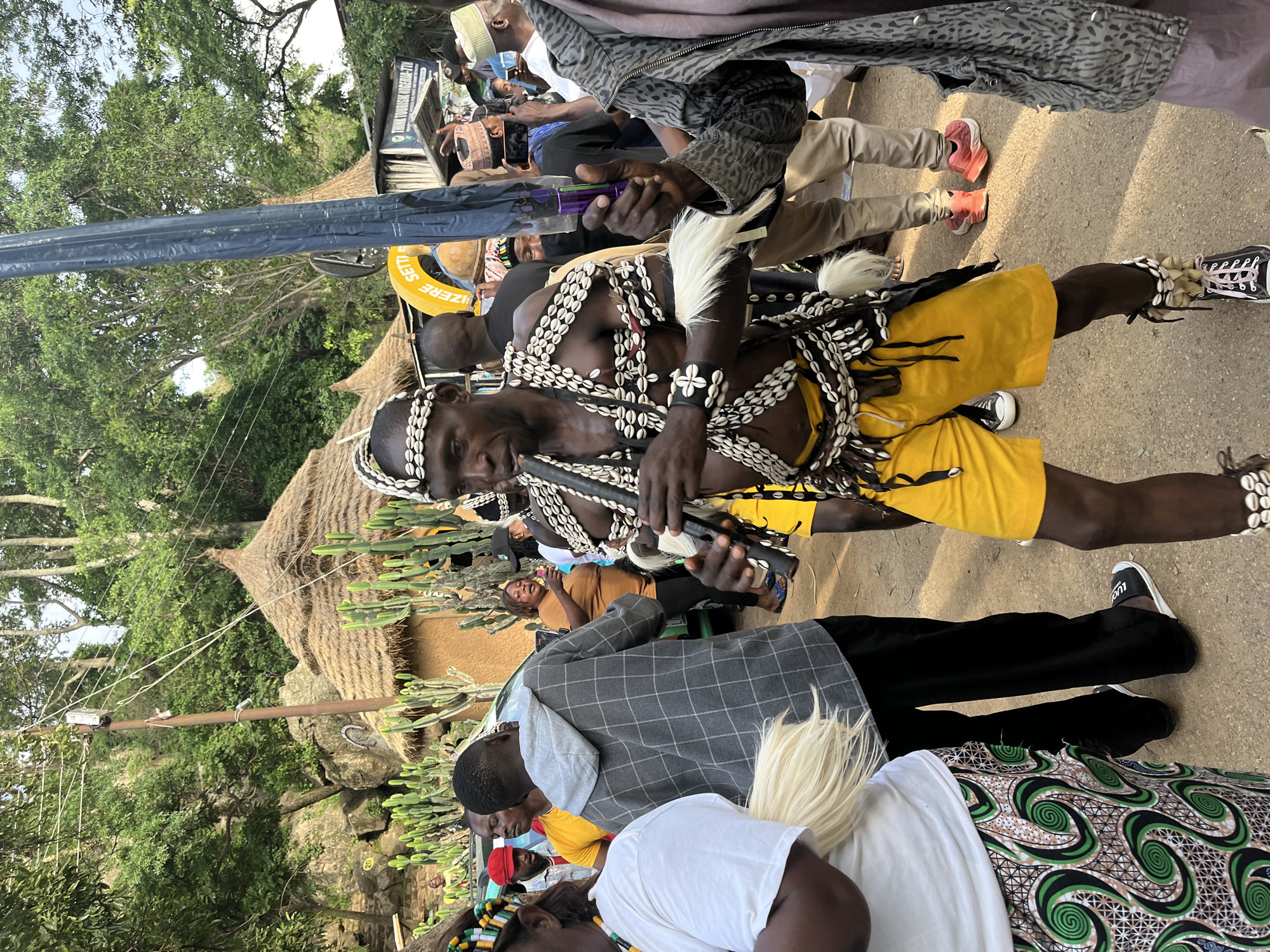 In Jos, Nigeria, a man in traditional attire plays a flute during a cultural event. He wears a beaded headdress and necklaces, a yellow loincloth, and leg adornments. He is surrounded by people, some of whom are taking photos. In the background, thatched-roof huts and cacti are visible. The scene appears to be outdoors, possibly at a festival or cultural gathering.