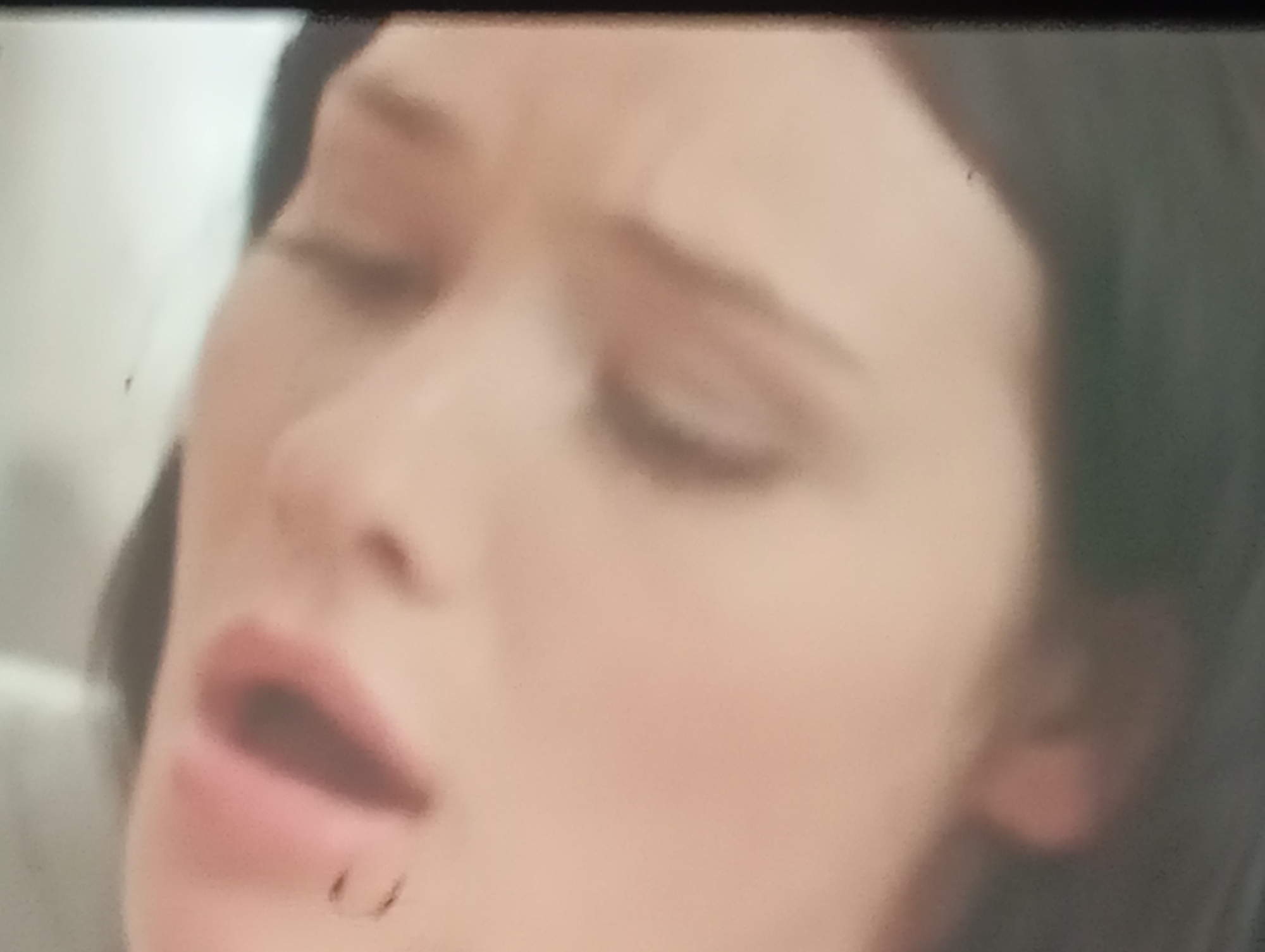 The image is a close-up, blurry, and low-resolution shot of a person's face. The person appears to be female, with fair skin and dark hair visible on the right side of the frame. Her eyes are closed, and her mouth is slightly open, revealing part of her teeth. There is a small, dark mark on her lower lip, which could be a mole or a blemish. The lighting is soft, with some areas appearing brighter than others, suggesting an indoor setting. The background is out of focus and appears to be a light color, possibly a wall or a curtain. No other individuals or objects are clearly discernible. The scene is devoid of any context that would link it to Sofiivska Borshchahivka, Ukraine, or any specific location.