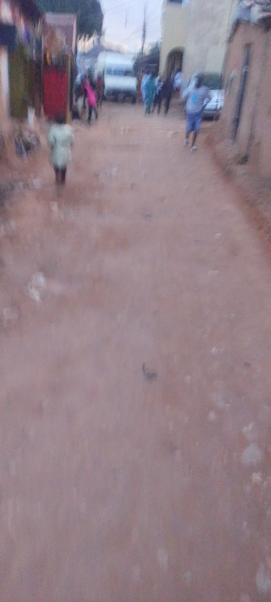 The image depicts a dusty, unpaved street in Gura Top, Nigeria, with several individuals present. In the foreground, a child wearing a light-colored garment is walking away from the camera. Further down the street, a white van is parked, and a group of people, some wearing blue and green garments, are gathered. Another person, wearing a grey t-shirt and blue shorts, is visible walking on the right side of the street. Buildings, appearing to be made of mud or brick, line the sides of the street, with some structures displaying red and green elements. The overall scene suggests a casual daytime activity in a residential or semi-commercial area.