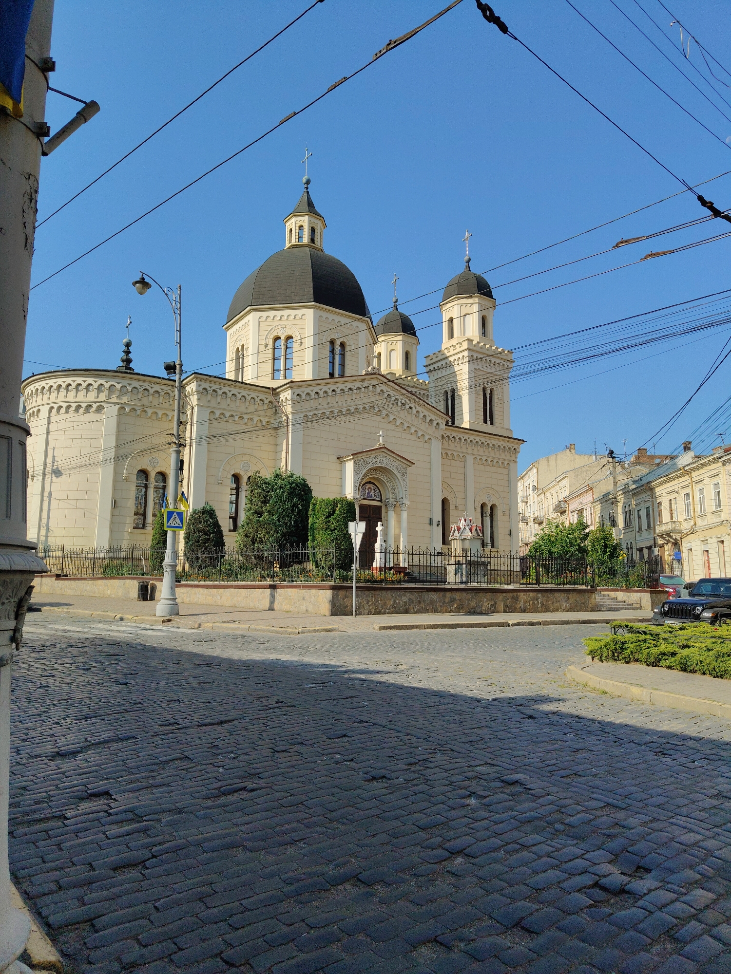 The image depicts the Holy Spirit Cathedral in Chernivtsi, Ukraine, on a clear, sunny day. The cathedral, a cream-colored building with a prominent black dome and two smaller domes, is the central focus. It features intricate architectural details, including arched windows and decorative cornices. In front of the cathedral, a black metal fence encloses a garden with neatly trimmed bushes and trees.
The street in front of the cathedral is paved with dark cobblestones, showing signs of wear and tear in places. A lamppost stands near the sidewalk, and a pedestrian crossing is visible. To the right of the cathedral, a row of older buildings with traditional European architecture lines the street, some with balconies and awnings. A black Jeep is parked on the right side of the street, near a patch of green landscaping.
The sky is a vibrant, cloudless blue, and overhead, numerous electrical wires crisscross the frame, suggesting an urban environment. A portion of a white pillar, possibly part of a street decoration or building facade, is visible on the far left of the image. The overall scene conveys a sense of calm and historical significance within a city setting.