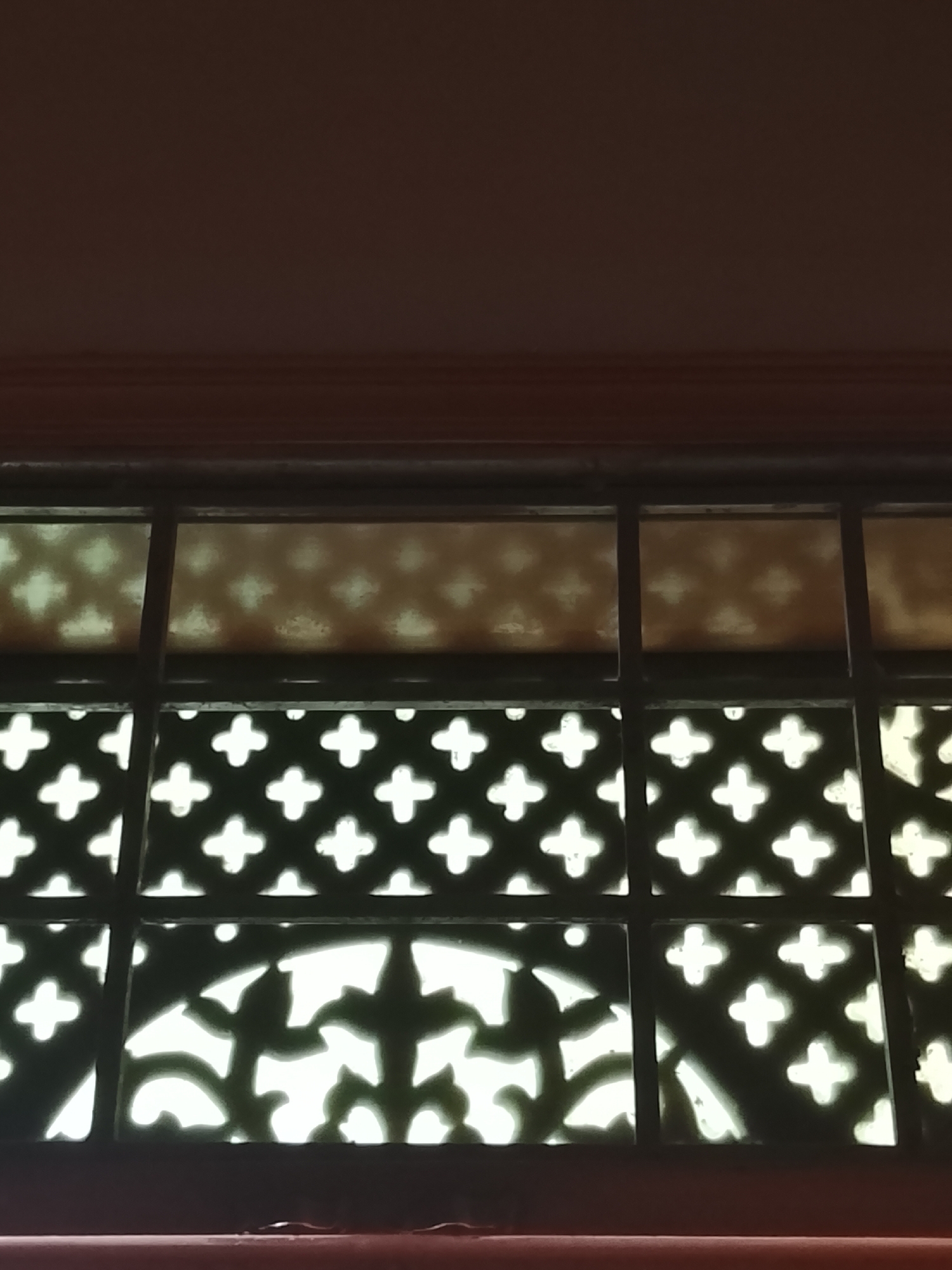 A partial view of an ornate metal window grille with geometric and floral patterns is depicted. The grill is dark and appears to be in front of a light source, causing the cut-out designs to appear bright. The grille is set in a dark-colored frame. Above the grille are additional window panes with a faint geometric pattern. The location details are not specified or clear in the image.