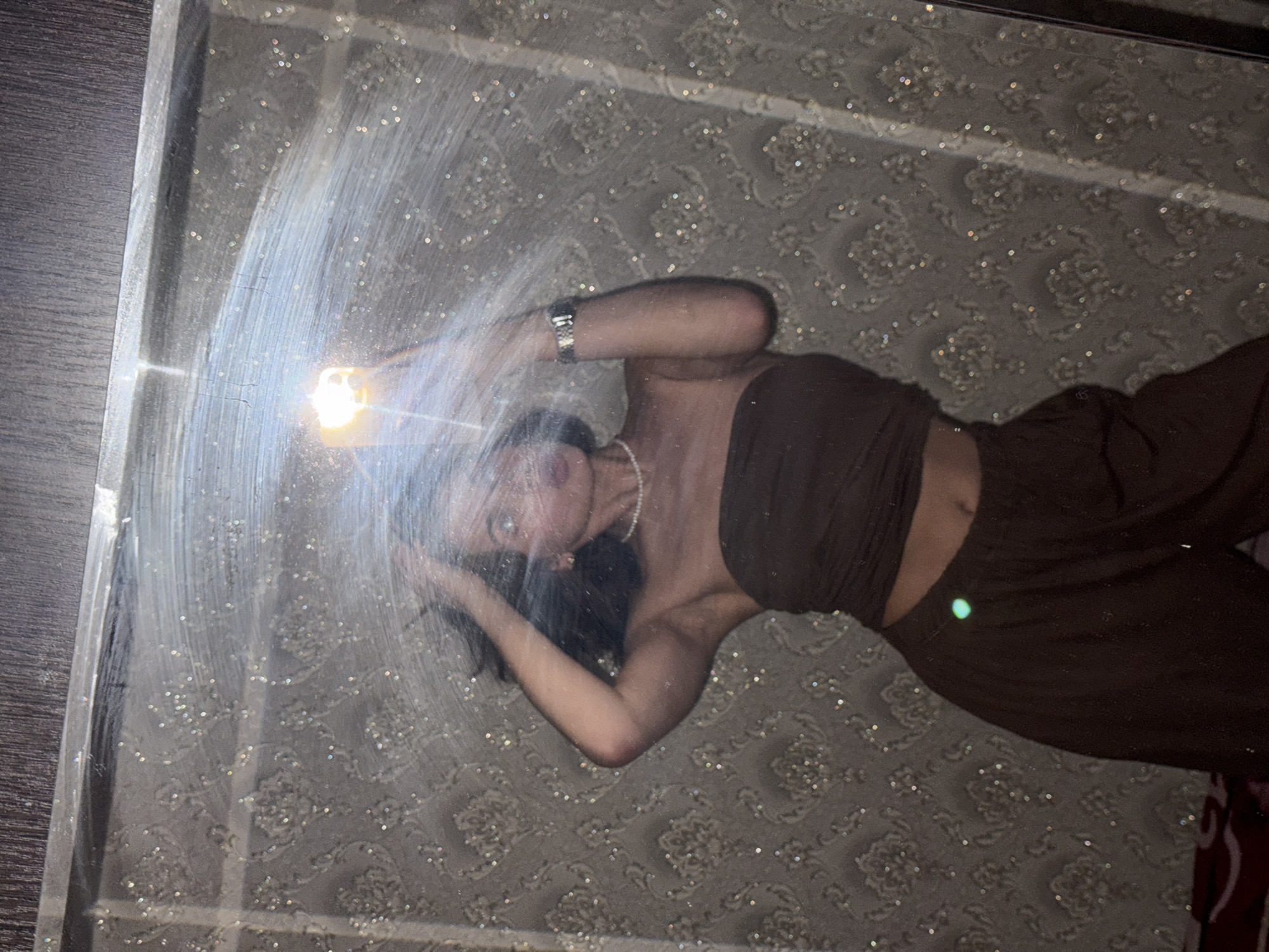 A woman wearing a brown bandeau top and matching pants lies on a rug with a floral pattern visible through a clear surface (possibly a glass table or a framed mirror). She wears a watch on her left wrist and a pearl necklace. Her left arm is raised, holding a light source that casts a bright glare. The scene is confined within a rectangular border, suggesting it is a photograph of a photograph. The location is not discernable from the provided image.