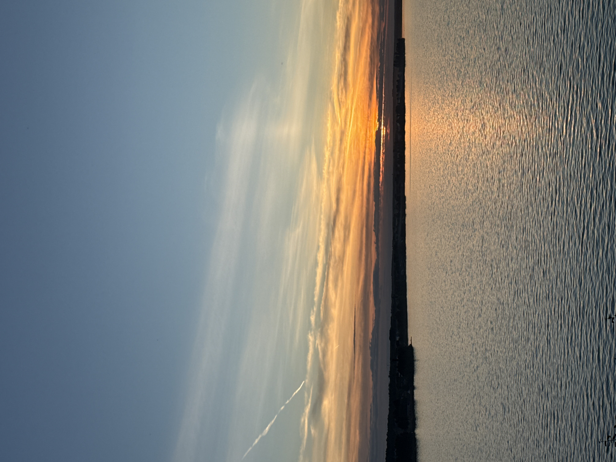 This is a vertical, high-angle, eye-level shot of a body of water, likely the Votkinsk Reservoir, at sunset. The sky transitions from a clear, light blue at the top to warmer hues of orange, yellow, and pink as it nears the horizon, where the sun is partially obscured by a dark band of clouds or land. Wispy clouds streak across the sky, with a prominent condensation trail from an aircraft visible in the upper left quadrant. The water is calm, with gentle ripples reflecting the colors of the sunset. A dark, silhouetted shoreline with indistinct structures is visible in the distance. The overall scene is serene and natural.