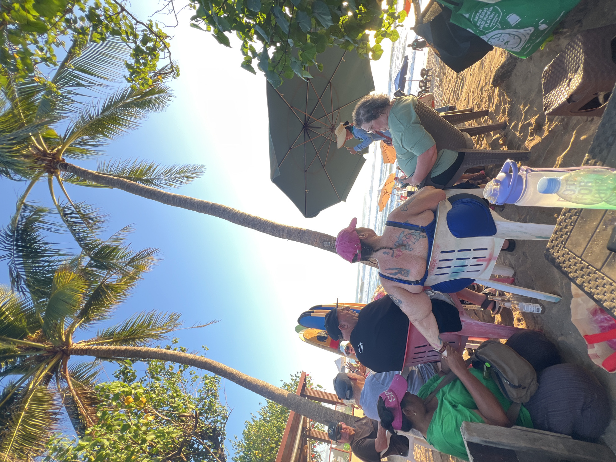 The image depicts a group of people sitting on a beach in Kuta, Indonesia, near palm trees and surfboards. The scene is set during the daytime with a clear blue sky. In the foreground, several individuals are seated on chairs, some facing away from the camera, observing the ocean. One woman in a green shirt is focused on her hands, while others are engaged in conversation or looking towards the water. In the background, beach umbrellas and lounge chairs are visible, and a few people are walking along the shoreline. The atmosphere suggests a relaxed, beachside gathering.