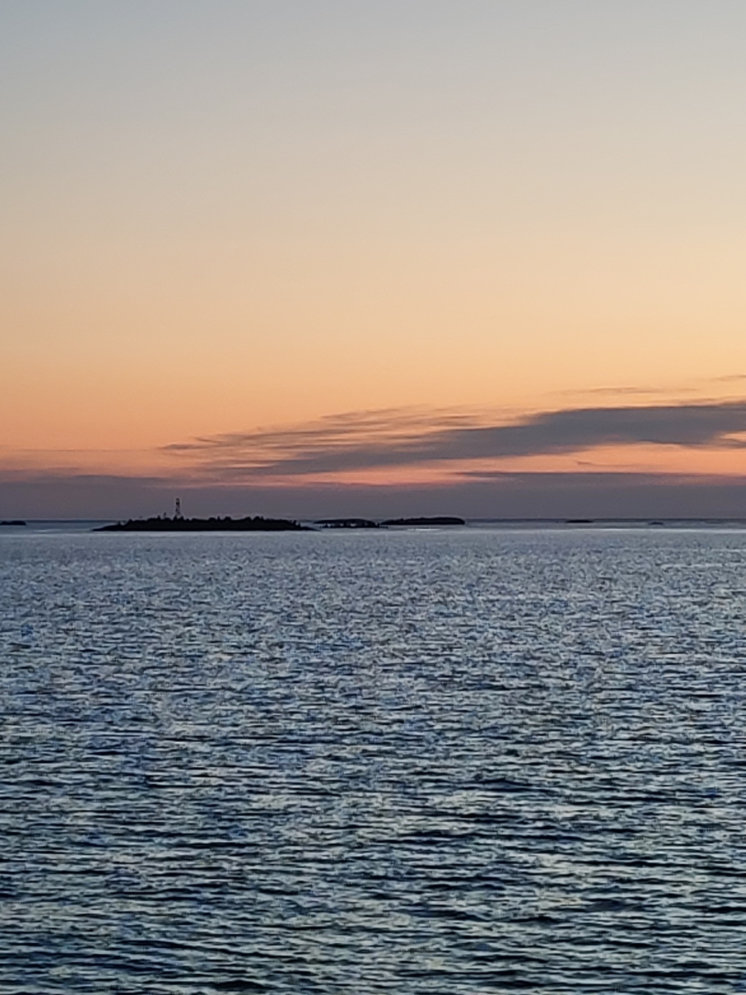 The image depicts a tranquil ocean scene at dusk, with the sky transitioning through shades of pale blue and soft orange. In the distance, a small, dark island is visible, dotted with what appears to be a lighthouse and scattered structures. The water in the foreground is calm, with gentle ripples reflecting the muted colors of the sunset. The overall atmosphere is serene and peaceful, suggesting a remote coastal location. The image does not provide any specific clues about the city of Shal'skii, Russia, as it appears to be a general seascape.