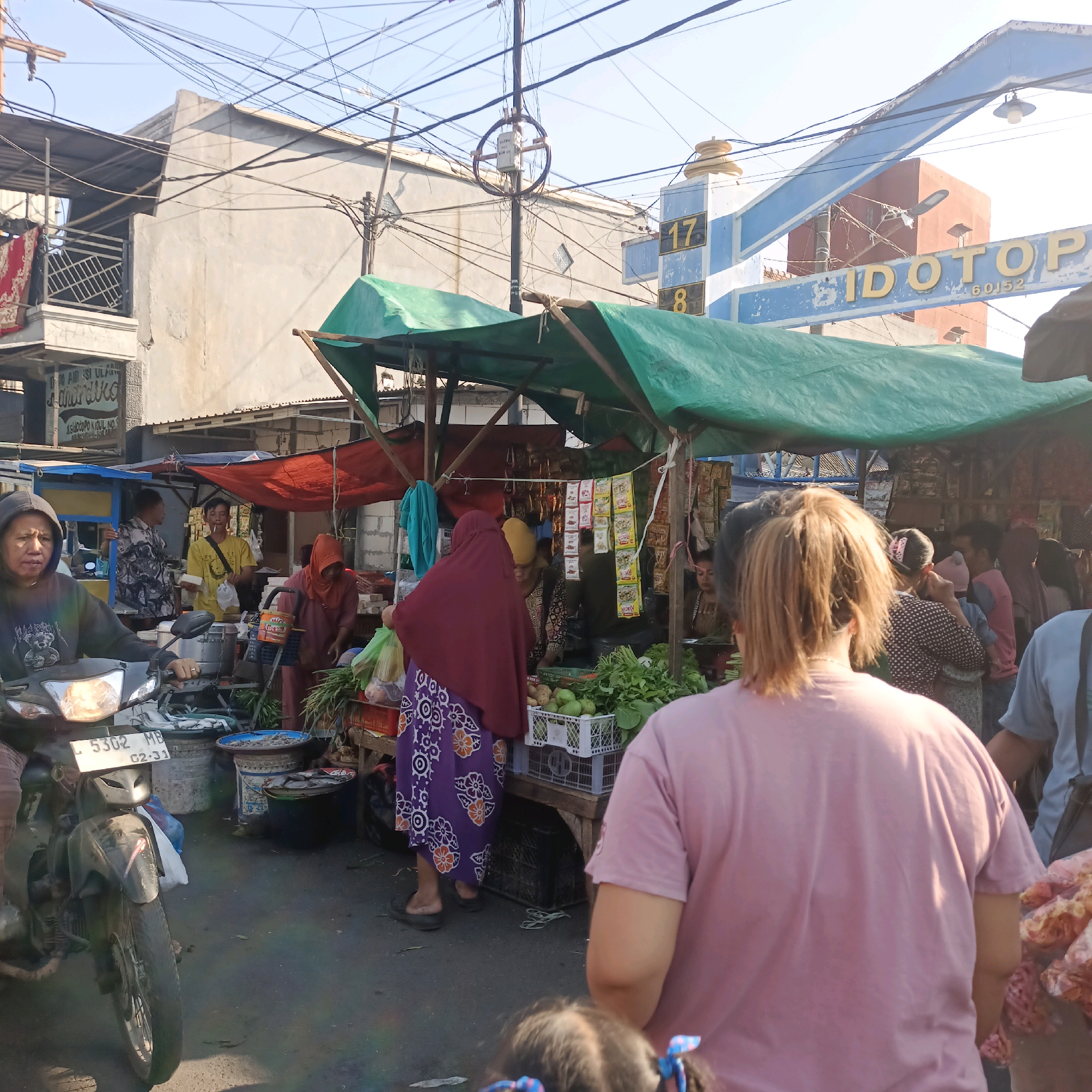 The image depicts a bustling street market scene in Surabaya, Indonesia, likely in a commercial area given the signage visible. In the foreground, a person is seated on a dark-colored motorcycle, facing the viewer's right, with their head and shoulders visible. The motorcycle has a license plate with the characters "L 5302 MB" and "02.31". The street itself is paved and appears well-trafficked.
Behind the motorcycle, a market stall is set up under a green canopy, displaying various produce including leafy greens, what appear to be guavas, and potatoes. Several individuals are engaged in market activities. A person in a maroon hijab and a purple patterned skirt is interacting with the stall, holding a plastic bag of produce. To their left, another individual in a red hijab is also present.
Further into the market, under a red canopy, a man in a yellow shirt is seen smoking and holding what looks like food. Another man in a patterned shirt stands behind a food cart. The background reveals buildings, including one with a sign that appears to read "IDOTO" and "60152", and an arched structure with the number "17". The sky is clear and bright, suggesting it is daytime. Electrical wires crisscross above the scene, a common feature in urban Indonesian environments. The overall impression is one of a lively and active marketplace.