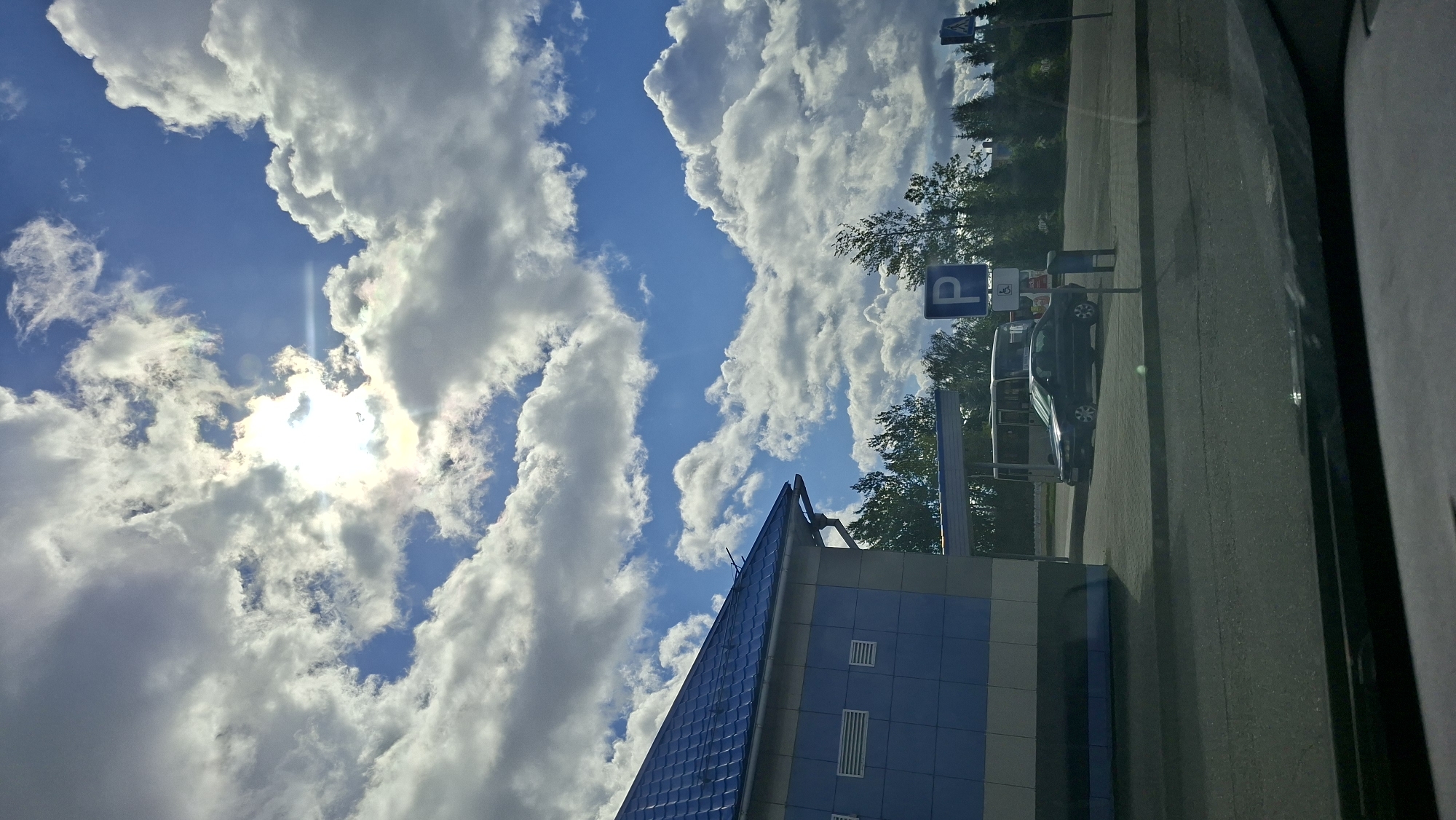 The image shows a view from inside a vehicle looking out at a gas station or rest stop in Myadzyel, Belarus. The sky is bright blue with large, fluffy white clouds, and the sun is partially obscured by clouds, creating a lens flare.
In the foreground, the dashboard of the vehicle is visible. Beyond the vehicle, there is a paved area with asphalt roads and a cobblestone walkway. To the left, a light blue building with a dark gray base and a blue tiled roof is partially visible. The building has horizontal white ventilation grilles.
In the middle ground, there is a black sedan parked on the cobblestone area. Behind the sedan, there is a bus parked under a canopy. A blue sign with a white "P" indicates parking, and next to it, a blue and white sign depicts a person in a wheelchair, indicating accessible parking. Further back, a line of green trees forms the background, and beyond that, more trees and some buildings are visible. A pedestrian crossing sign is visible on the right side of the frame. The overall scene suggests a typical roadside service area on a cloudy but bright day.