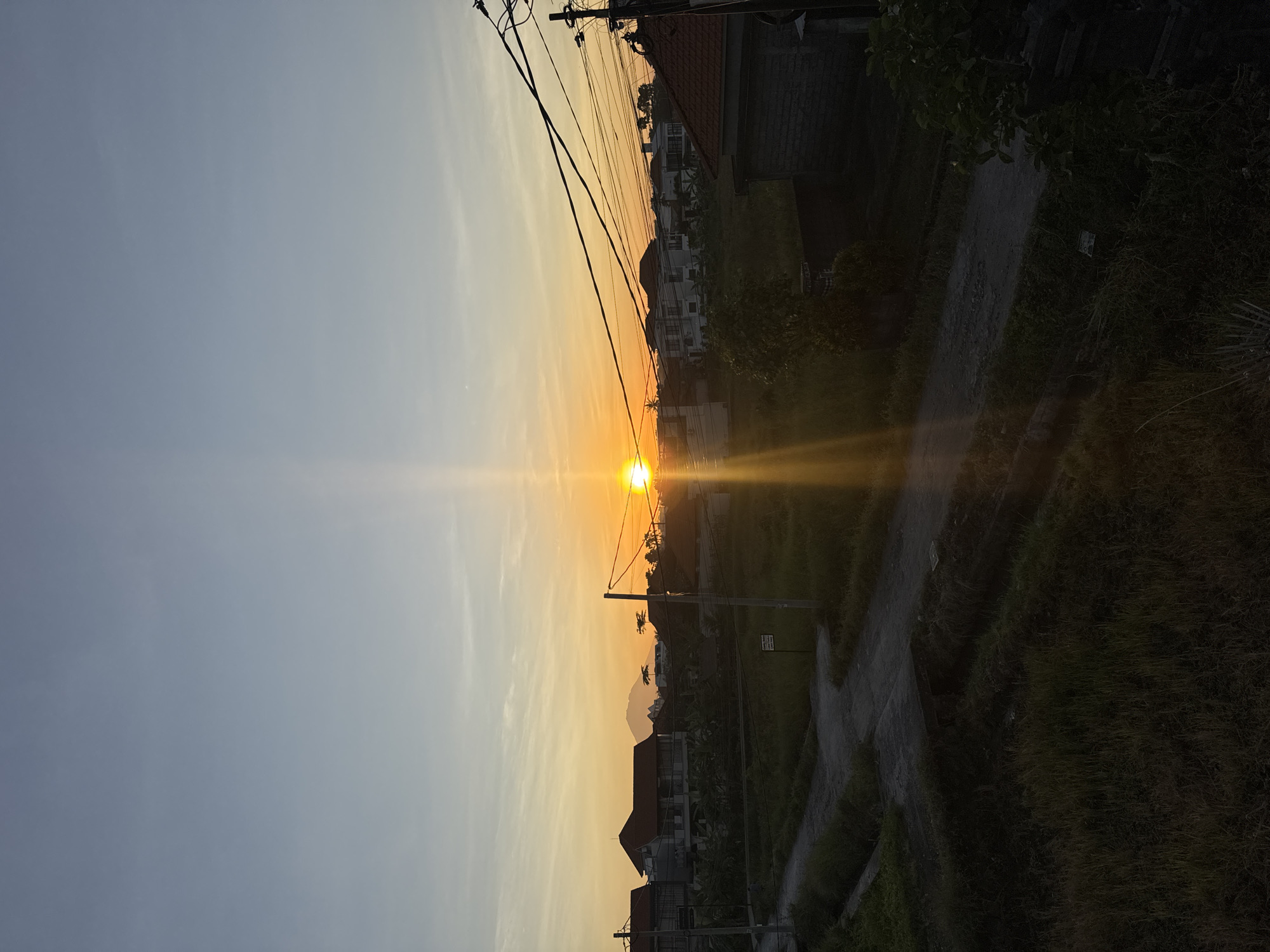 The image depicts a sunset in Mengwi, Indonesia. A bright, setting sun is positioned between buildings and electrical wires, creating a radiant glow. A road runs along the right side of the frame, bordered by vegetation. Buildings of varied heights are visible on both sides of the road, with some appearing dark due to the backlight. The sky transitions from a pale yellow near the horizon to a muted blue higher up. A mountain silhouette is visible in the distance, adding depth to the composition.