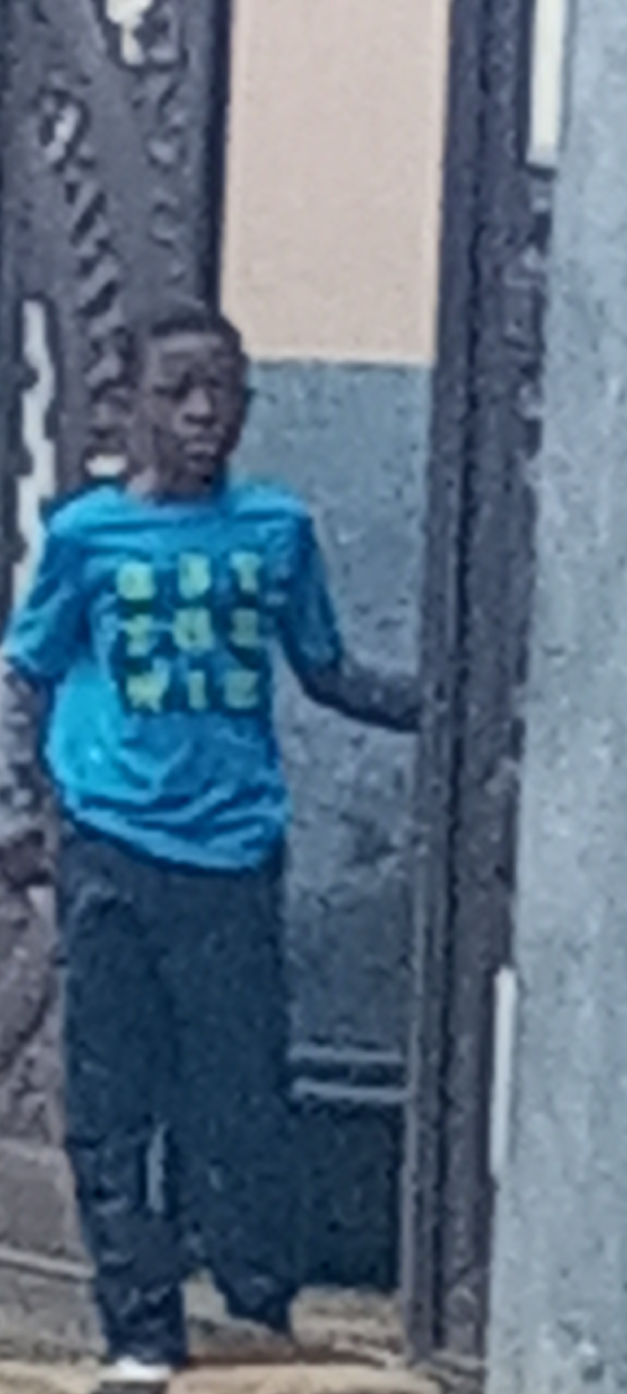 A youth stands next to a grey building with a light tan section visible above his head. The youth is wearing a bright blue short-sleeved t-shirt with green lettering and dark-colored pants. Part of a metal design is visible to the left of the youth. The location is Sabon Tasha, Nigeria.