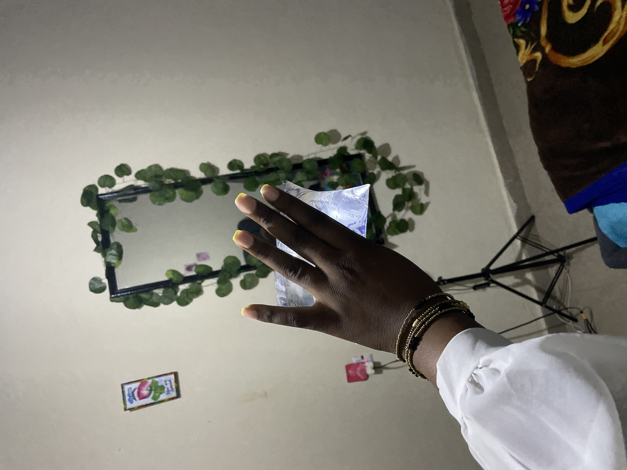 A close-up of a person's left hand is held in front of a wall, with a mirror and some decorative plants in the background. The hand has dark skin and is adorned with several beaded bracelets around the wrist and painted fingernails with yellow tips. The person is wearing a white, long-sleeved blouse. The hand is holding a clear plastic bag that appears to contain water or a clear liquid, and the bag has some text and markings on it. To the left of the hand, a pink electrical outlet is visible on the wall. Behind the hand, a black tripod stand is positioned, and further back, a rectangular mirror is decorated with artificial green vines. The wall is painted a neutral, light color. The floor visible at the bottom of the image is covered with a brown blanket featuring a floral pattern. The scene suggests an indoor setting, possibly a bedroom or a personal space. The location is identified as Rafin Par, Nigeria.