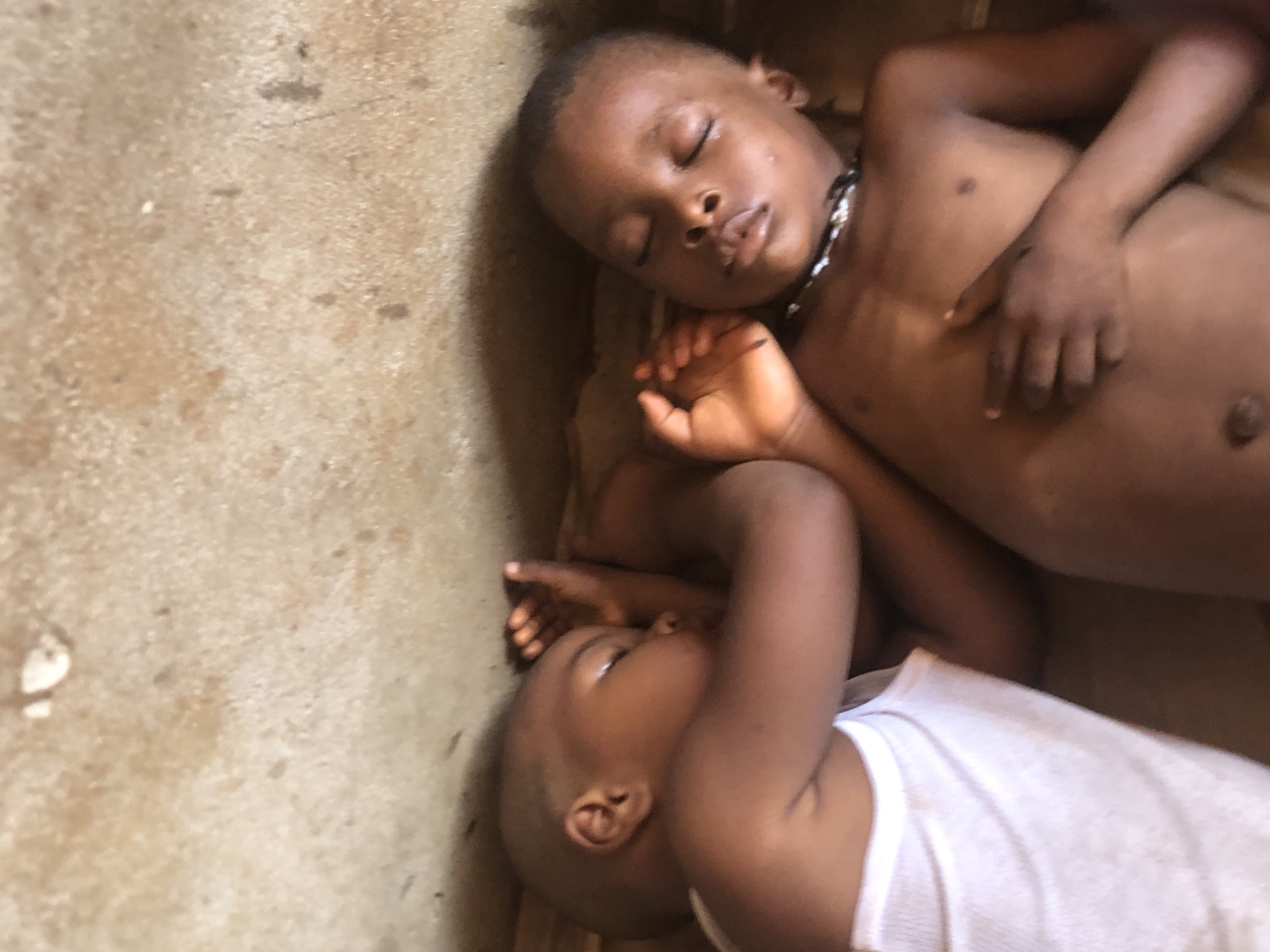Two young children are sleeping on a dirt floor. The child in the foreground is lying on their side, wearing a white tank top, with their arm bent and hand near their face. The child in the background is lying on their back, with their eyes closed and wearing a dark necklace. The background is a rough, textured dirt surface. The scene appears to be an outdoor or very basic indoor setting given the dirt floor. The lighting suggests it could be daytime, but no specific time can be determined. The overall mood is one of peaceful rest. There is no visible text in the image.
