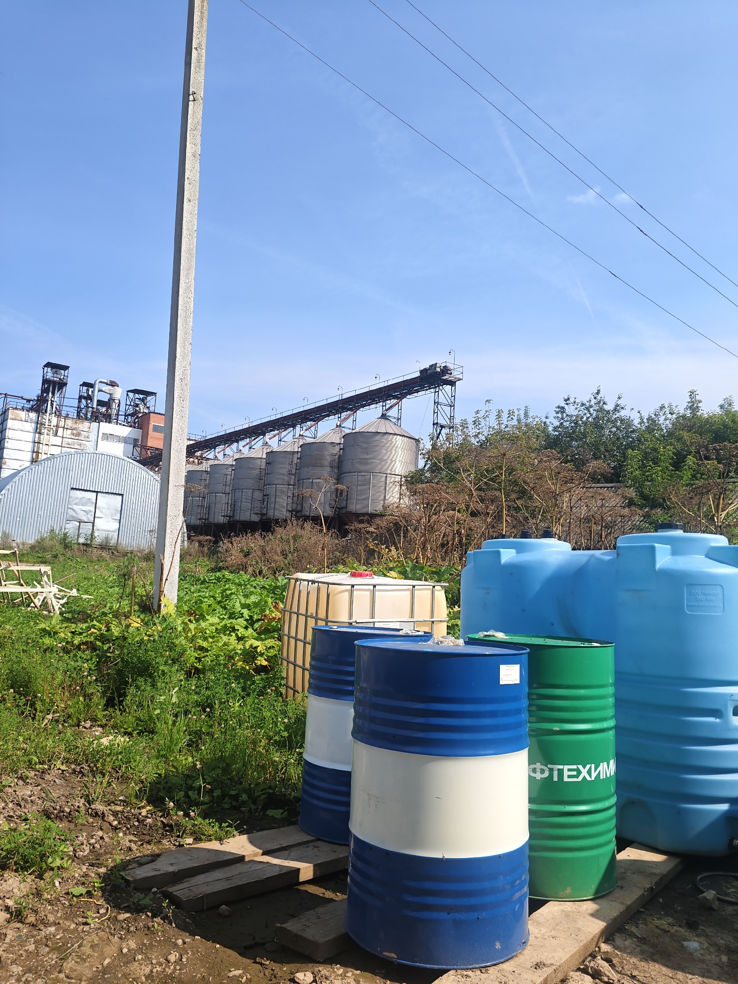 The image shows an outdoor industrial or agricultural scene under a clear blue sky with some thin clouds and power lines. In the foreground, there are several large barrels and water tanks. To the left, a concrete utility pole stands tall. Behind the barrels, a large, beige IBC tote is visible. Further back, a row of cylindrical metal silos and industrial buildings are prominent, suggesting a facility for storage or processing. Lush green vegetation and dry, tall plants fill the midground. The overall impression is one of an operational industrial site in a rural or semi-rural environment. The presence of the industrial structures and the general landscape suggests this may be in the vicinity of Платошино, Russia, known for its agricultural and industrial activities. There are no people or animals visible in the image.