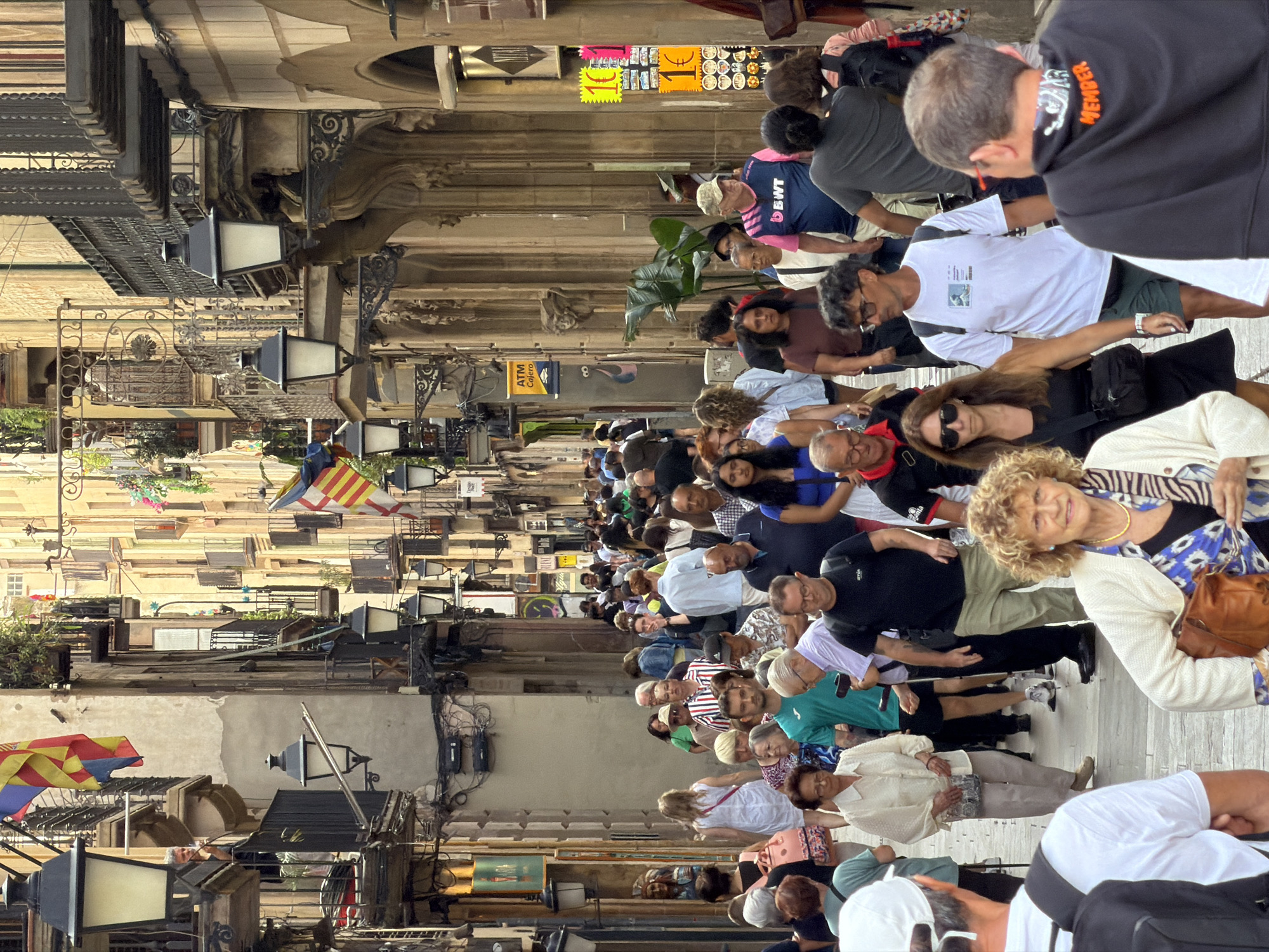 The image depicts a crowded street scene in what appears to be a European city, with the location context indicating Barcelona, Spain. A large group of people are walking down a narrow street lined with buildings. The buildings have a traditional architectural style with balconies and ornate detailing. Several lampposts are visible along the street.
The people are diverse in age and are dressed in casual clothing, suggesting it is daytime and likely warm weather. Some individuals are looking at their phones, while others are engaged in conversation. There's a general sense of movement and activity, possibly indicating a tourist area or a marketplace.
In the background, several signs are visible. One sign, in yellow and black, has the currency symbol "€" and the number "10" on it, suggesting a price. Another sign reads "ATM Caixero". There is also a sign with the text "MEMBER" on the back of a black hoodie. Flags, one of which appears to be the Catalan flag, are also visible, adding to the location context. The overall mood seems lively and bustling.