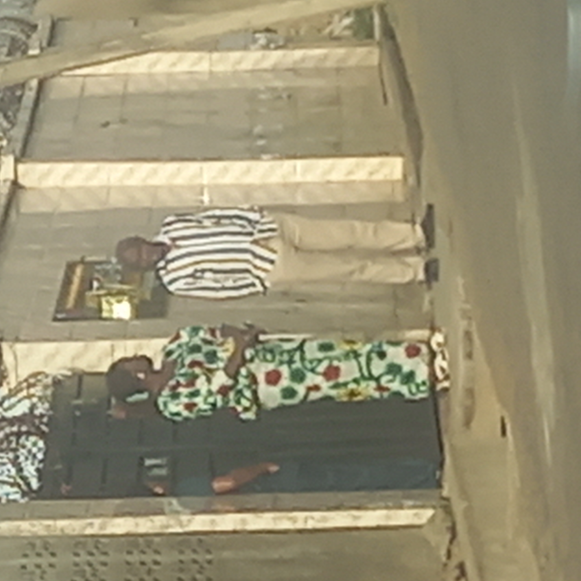 The image, taken in Barden Yana, Nigeria, captures an overhead view of at least three individuals resting on a tiled surface during the daytime.
In the upper section of the frame, a person is lying on their back, wearing a white shirt with brown/gold and black stripes and light-colored trousers. Their head is towards the left. Near their head, on a small, dark, rectangular mat, are a small framed object (possibly a picture) and some shiny, golden-hued items, which could be personal belongings or offerings.
Below this individual, another person is lying down, also on their back with their head oriented to the left. They are dressed in a brightly patterned garment featuring green, red, and yellow motifs on a white background.
Further down, a third person is partially visible, mostly covered by a dark mat or blanket. Only their lower legs and one bare foot are clearly seen. All three individuals appear to be resting or sleeping.
The ground consists of a light-colored tiled surface with rectangular patterns and darker grout lines or strips. A light-colored vertical structure, possibly a wall or pillar, is visible on the right side of the image. The even lighting suggests it is daytime, possibly under an overcast sky or during early morning or late afternoon, as there are no strong, defined shadows. No visible text is present in the image.