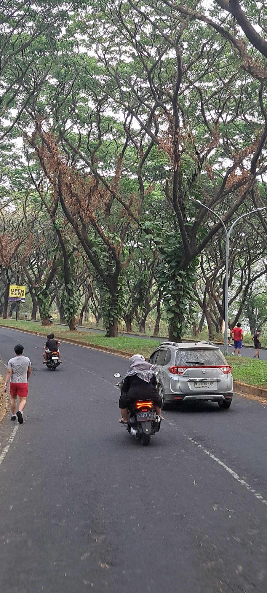 The image depicts a tree-lined road in Bulukandang, Indonesia. In the foreground, a person walks away from the camera on the left side of the road, wearing a gray t-shirt and red shorts. Further ahead on the road, a motorcycle with two occupants is seen from behind, with the passenger wearing a dark top and headscarf. To the right, a silver SUV is driving on the road. The environment features numerous large trees with extensive branches and green foliage, with some dried brown branches visible. There are also streetlights and a yellow "OPEN HOUSE" sign visible on the side of the road in the distance. The road appears to be asphalt, with a white line marking the lane. In the background, other people can be seen walking or standing on the grassy embankment.