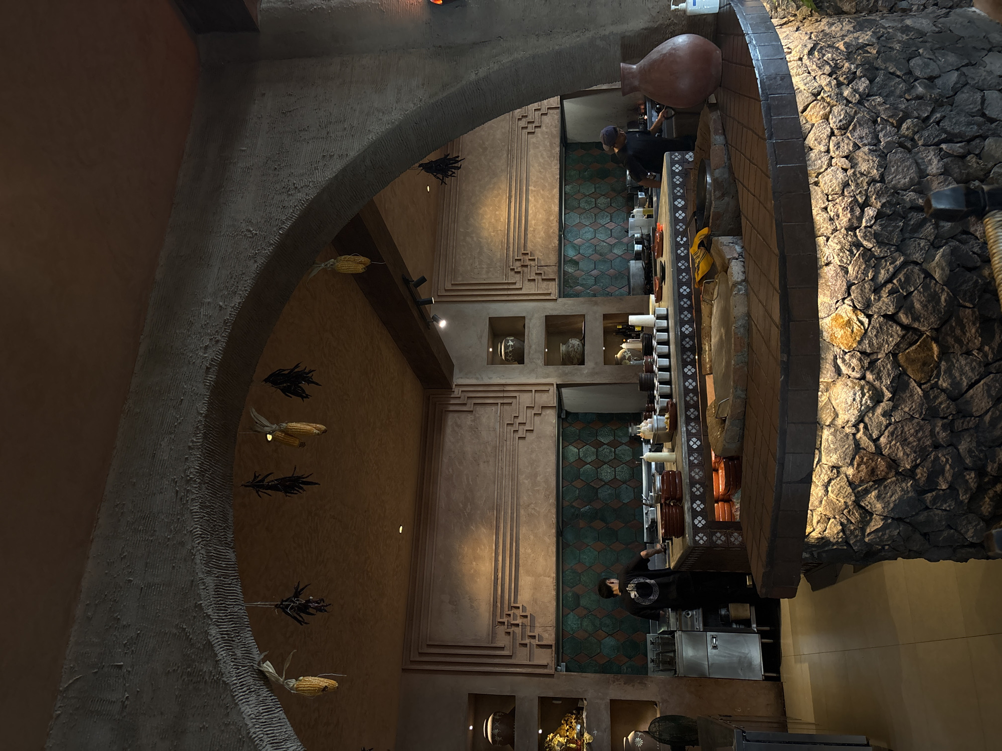 Captured from an elevated, slightly skewed perspective, the image portrays the interior of a building. An archway adorned with decorative corn and plant stalks dominates the left side of the frame. The wall inside the archway is paneled with stepped decorative molding. The floor is tiled in a geometric pattern in green and brown. Within the building, at least two individuals are visible, possibly interacting with a serving station or preparation area filled with various containers, equipment, and food items. A stone wall with plants on top flanks the right side of the frame. The scene conveys an atmosphere of a restaurant or food preparation area. This is located in Mengwi, Indonesia.