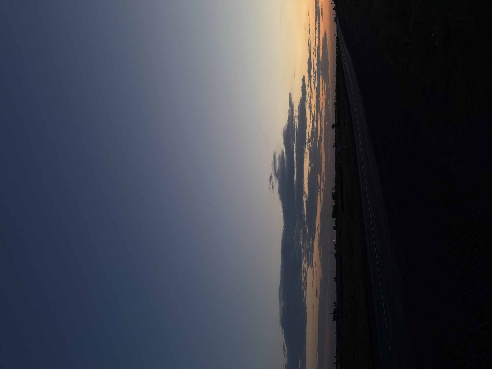 The image depicts a landscape at dusk or dawn. A two-lane road stretches into the distance along the right side of the frame. The surrounding landscape appears to be fields, transitioning from lighter tones near the horizon to dark areas at the right edge of the image. The sky dominates the left side of the image, ranging from a dark blue-gray at the bottom to a lighter, gradient color transition toward the horizon where the sky is touched with light orange hues. Several clouds are present near the horizon, showing darker areas of gray and silhouettes of trees or buildings, possibly power lines in the distance, are visible along the horizon line. The location is an unidentified rural area.
