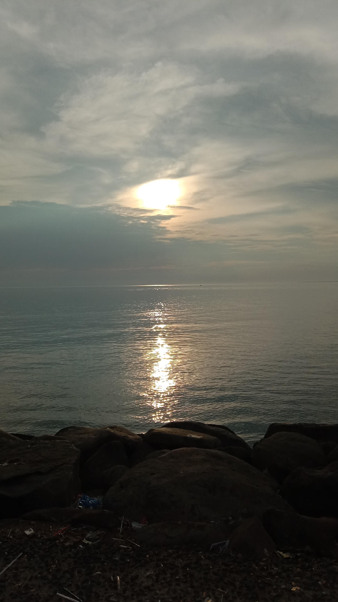 The image depicts a seascape at sunset, likely near Lhokseumawe, Indonesia. The sky is overcast with a bright sun partially obscured by clouds, casting a vertical column of light across the water's surface. Below the horizon line, the water is calm with a slight ripple texture. The foreground is composed of dark, uneven rocks that appear to form a breakwater or shoreline. Debris and trash are scattered on the ground among the rocks. There are no people visible in the image.