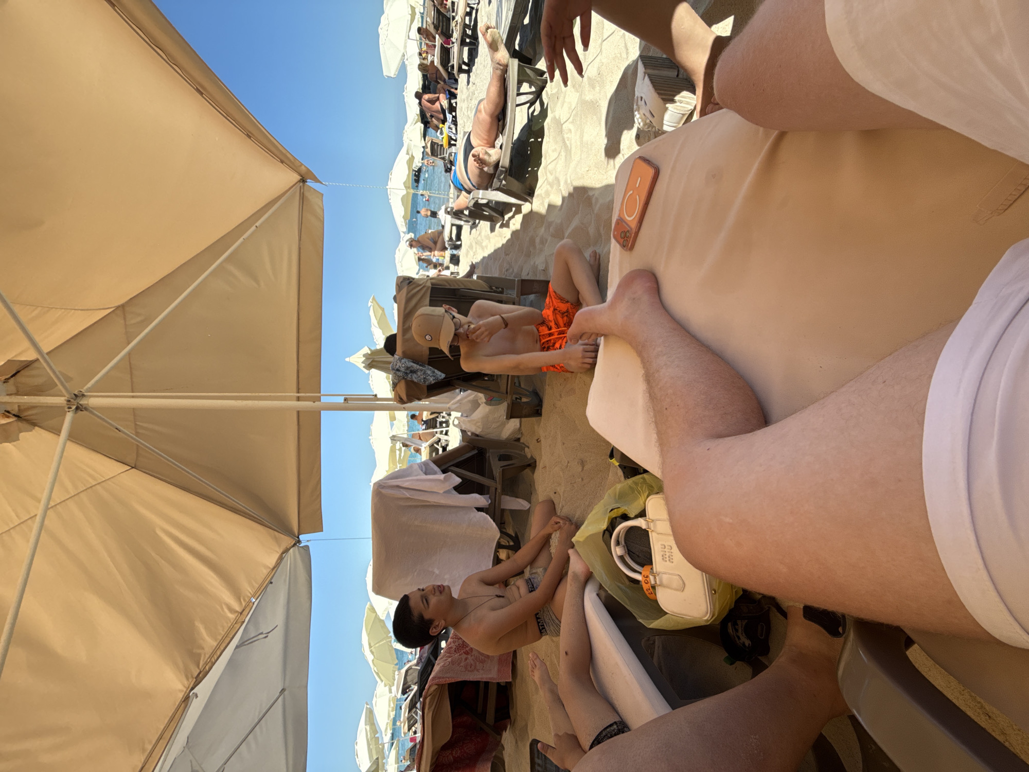 The image depicts a sunny day at a crowded beach. The foreground is dominated by the legs of several individuals, indicating a close-up perspective from someone sitting or lying down on a beach chair or the sand. Beyond them, a young boy wearing an orange swimsuit and a baseball cap is seated on the sand, looking down and appearing to eat. To his right, a white handbag with "miu miu" visible on it rests on the sand. In the background, numerous beach umbrellas and loungers are occupied by people sunbathing and relaxing. The sea and a clear blue sky are visible in the distance, suggesting a warm, pleasant weather. The overall atmosphere seems relaxed and vacation-like. The time of day is clearly daytime, indicated by the bright sunlight and shadows. There is no discernible text other than the brand name on the handbag.